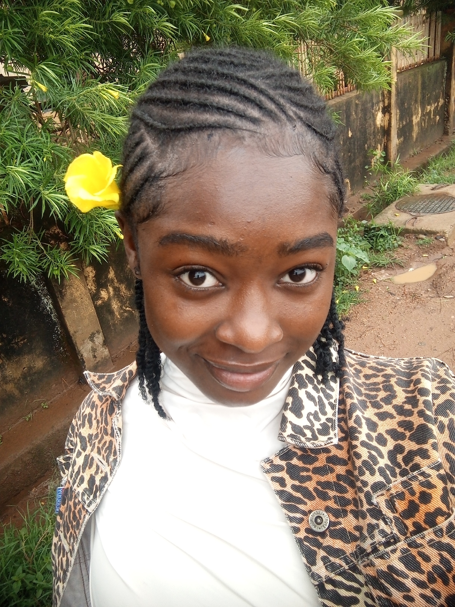 A young woman of African descent is captured in a close-up, frontal selfie. She has dark skin and her hair is styled in braids, with a single yellow flower tucked behind her left ear. She is wearing a white turtleneck shirt and a leopard-print jacket. The background suggests an outdoor urban environment, possibly in Rafin Par, Nigeria, with lush green foliage to the left and a weathered concrete wall and a dirt path to the right. The path shows signs of recent rain with puddles of water and a metal grate.