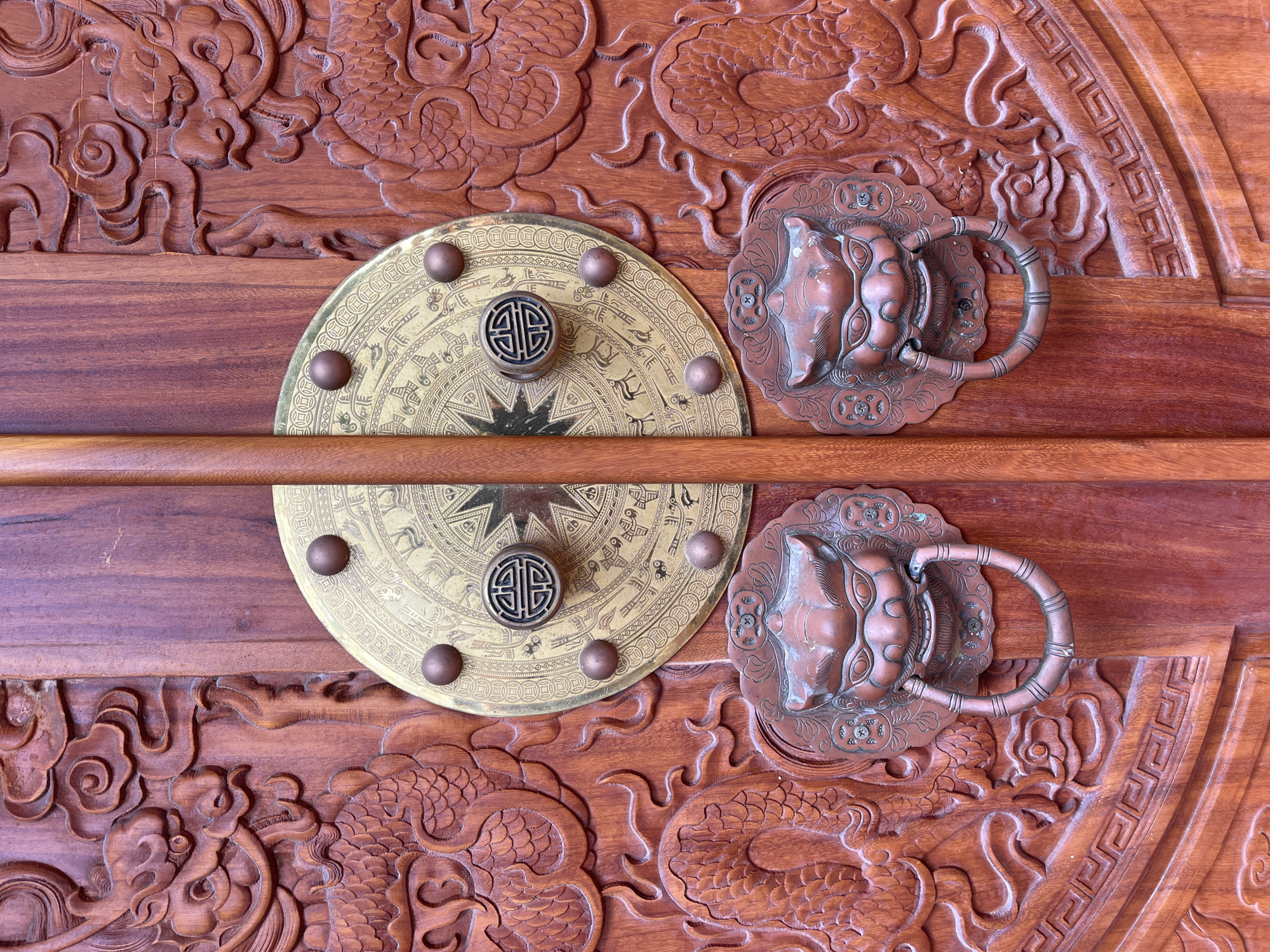A close-up, eye-level shot captures the intricate details of a traditional wooden door in Mũi Né, Vietnam. The doors are made of rich, reddish-brown wood and are heavily carved with Asian motifs. The left door features a detailed carving of a dragon with flowing clouds and scales, while the right door mirrors this design.
Dominating the center of the doors are two large, circular brass plates, each adorned with a star-like pattern and what appear to be astrological symbols or depictions of animals. Each brass plate has a prominent, dark, circular handle in the center, featuring a stylized knot or character design. Around the brass plates, several small, bronze-colored studs are visible.
Below each brass plate, a heavy, bronze lion-head door knocker is mounted. The lion heads have fierce expressions, with open mouths and ring handles attached. The entire surface of the doors is covered in these elaborate carvings and metalwork, suggesting a place of significant cultural or religious importance. Two vertical wooden door separators run down the center, obscuring a full view of the door seam. The lighting is even, highlighting the textures and depths of the carvings.
