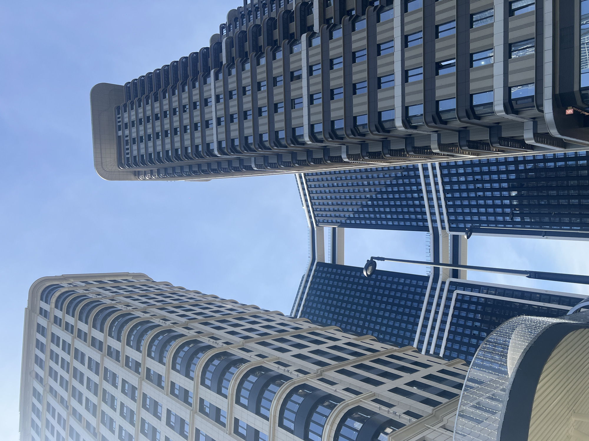 This low-angle photograph, taken in Khimki, Russia, captures a dynamic perspective of several modern high-rise buildings against a clear, bright blue sky. The image is rotated counter-clockwise, making the skyscrapers appear to lean dramatically across the frame.
In the foreground and to the left, a prominent light-colored building, possibly cream or light gray, displays a contemporary design with a smooth, rounded corner. Its facade is characterized by an alternating pattern of rectangular windows and distinctive arched window sections, complemented by vertical architectural lines. To the top and right, another towering structure with a darker gray and black color scheme features a more intricate, ridged design, incorporating numerous rectangular windows and noticeable vertical protrusions. In the mid-ground, further back, a large, dark blue or black building with a dense, grid-like pattern of small windows can be seen, interlinked by white or light-colored structural elements that form a bridge-like connection between its sections.
A street lamp or lamppost is visible in the middle-right, extending upwards, suggesting an urban streetscape. In the bottom right corner, a curved, decorative architectural element with a textured, possibly illuminated, surface is partially visible. The clear, cloudless sky indicates a sunny day. There are no people visible, allowing the focus to remain entirely on the impressive scale and architectural details of the buildings. A small red sign with the text "1/3" is faintly discernable on the darker, ridged building.