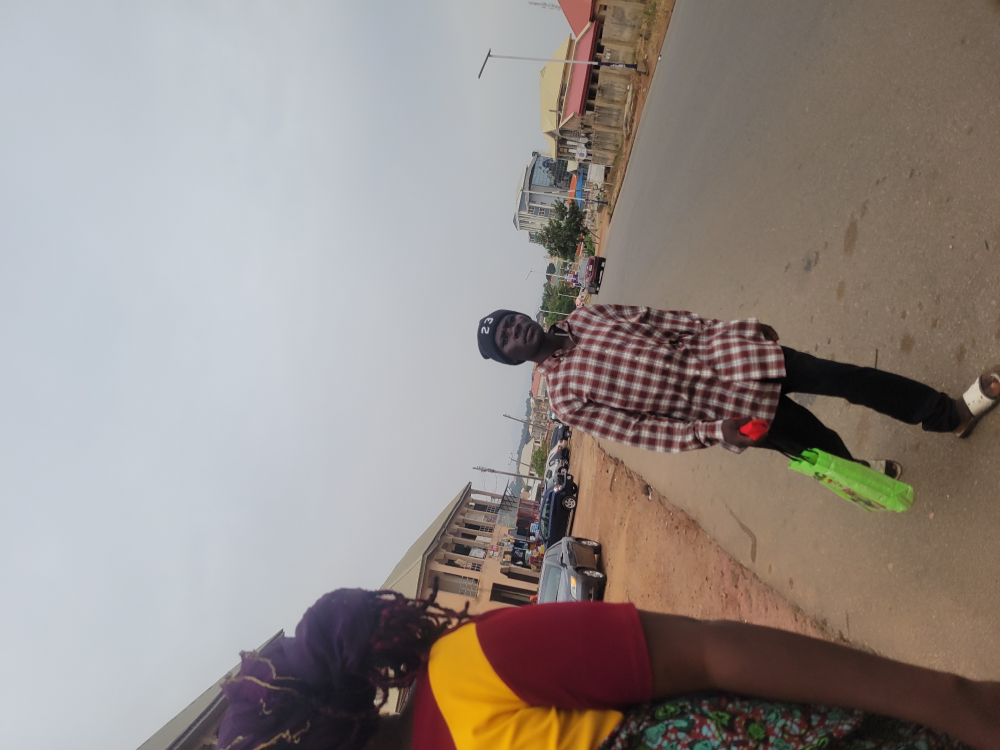 This image depicts an outdoor street scene in a town or city, likely in Jos, Nigeria, under an overcast sky. The perspective is slightly angled, showing a paved road extending into the distance, flanked by buildings and parked vehicles.
In the mid-ground, a person stands on the road's edge, looking directly at the camera. They are wearing a black beanie with the numbers "22" (or "Z2") prominently displayed, a long-sleeve shirt with a red and white plaid pattern, black trousers, and open-toed sandals that are white and brown. They hold a bright neon green bag in their left hand and a small red object in their right.
Partially visible in the foreground, with their back to the camera, is another person. They wear a purple head covering over dark braided hair, a top featuring red sleeves and a yellow back, and a lower garment (possibly a skirt or wrapper) with a multi-colored pattern including red, green, and blue. This person appears to be moving along the roadside.
The road surface is asphalt, showing some wear and scattered dirt, with a visible sandy or earthen shoulder. In the background, various buildings line the street, ranging from multi-story residential or commercial structures to one with a modern glass facade. Several cars are parked along the road, and street lights are spaced along the pavement. The sky is a uniform, light grey, indicating an overcast daytime with diffused light and no clear shadows.
