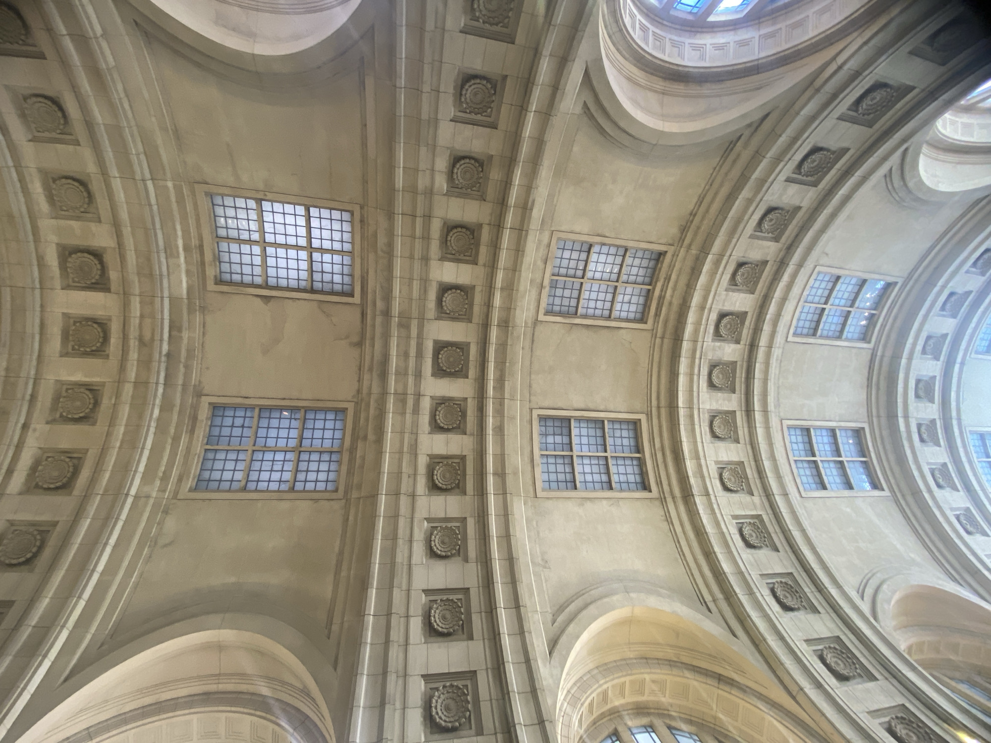 The image captures an upward view of the intricate ceiling of a building in Buenos Aires, Argentina, possibly a grand hall or public space. The ceiling features a symmetrical, arched design with repeating patterns of decorative rosettes and rectangular windows arranged in a grid. The structure is made of light-colored stone or plaster, which has an aged, slightly weathered appearance. The windows allow natural light to filter in, creating a soft, diffused illumination. The perspective is angled, suggesting the photographer was standing directly beneath the ceiling, emphasizing its height and grandeur.