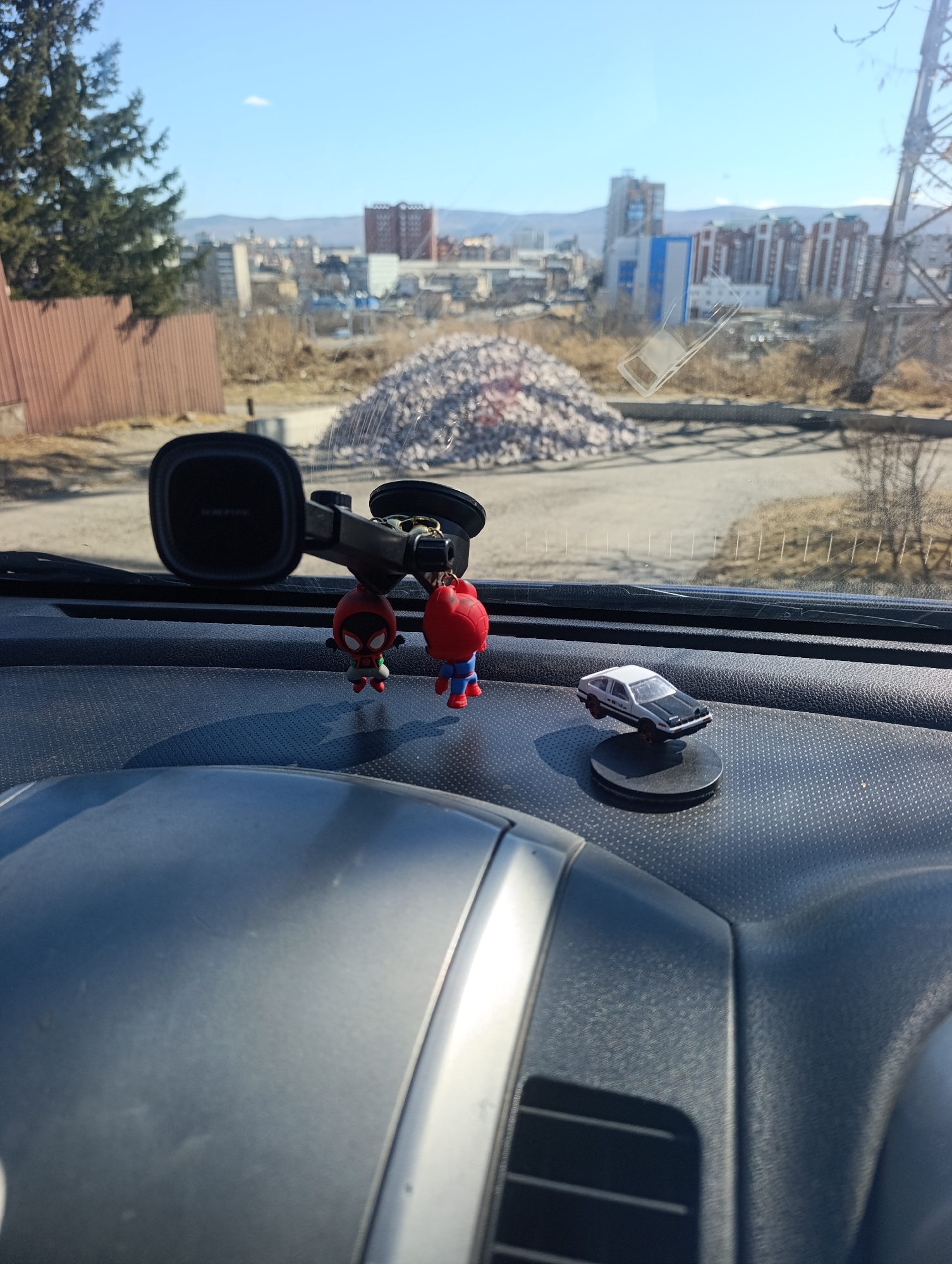 The image, likely taken from inside a car, shows a view of a cityscape in Krasnoyarsk, Russia. The car's dashboard is in the foreground, with a suction cup phone mount holding a couple of Spider-Man keychains. A small model of a black and white car is also on the dashboard. Through the windshield, a pile of rubble is visible, along with buildings of varying heights and architectural styles, typical of Krasnoyarsk. The sky is clear and blue. A partial view of a transmission tower is present to the right side of the image.