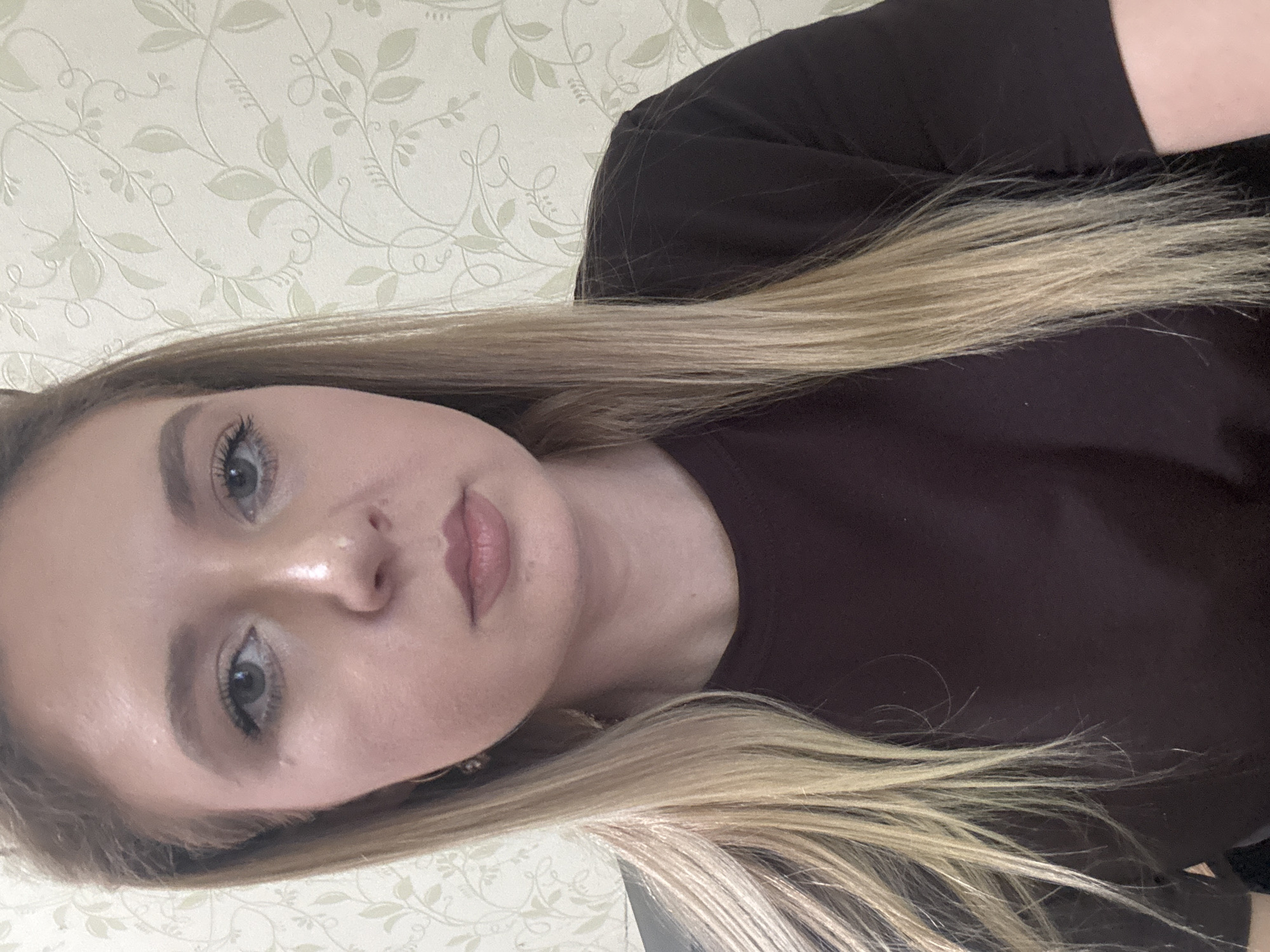 A young woman with blonde hair and blue eyes is pictured from the chest up. She is wearing a dark brown t-shirt and her hair is worn down. She is looking directly at the camera with a neutral expression. The background is a textured wallpaper with a light green floral pattern. The image appears to be a selfie taken indoors. The location is unknown.