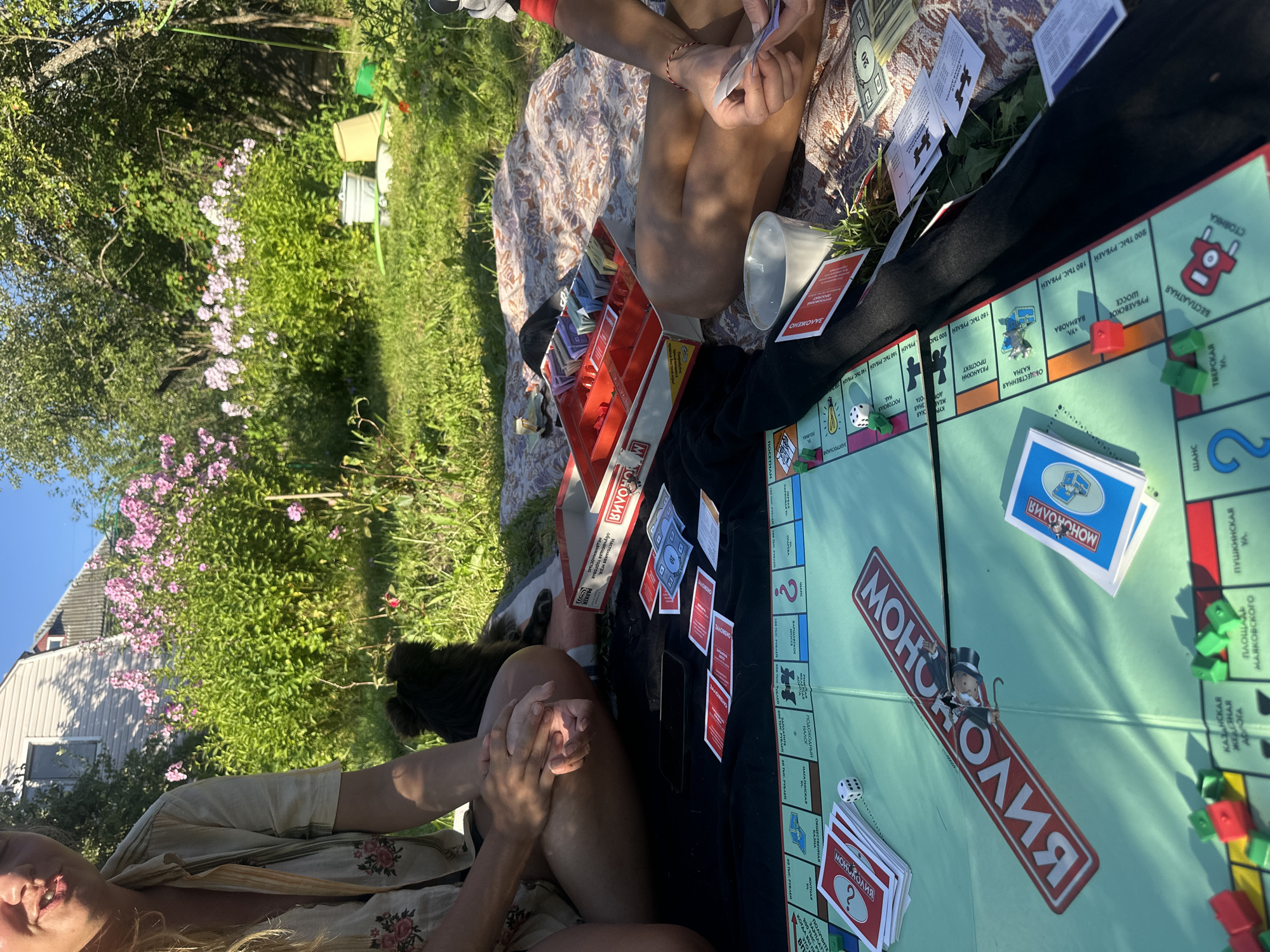 In the city of Krasnaya Presnya SNT, Russia, a group of people are enjoying a game of Monopoly on a sunny day. The game board is spread out on a patterned blanket on the grass, surrounded by green trees and pink flowers. Two individuals, with their legs visible, are seated on the blanket, engrossed in the game. One person is holding Monopoly money, while the other's hands are clasped. A small black dog is also present, sitting near the people. Scattered around the game board are Monopoly cards, dice, and plastic houses and hotels. A red Monopoly box, filled with game pieces and money, sits to the side. The scene is set in a backyard or garden, with a house visible in the background. The lighting suggests it is daytime, with the sun casting shadows.