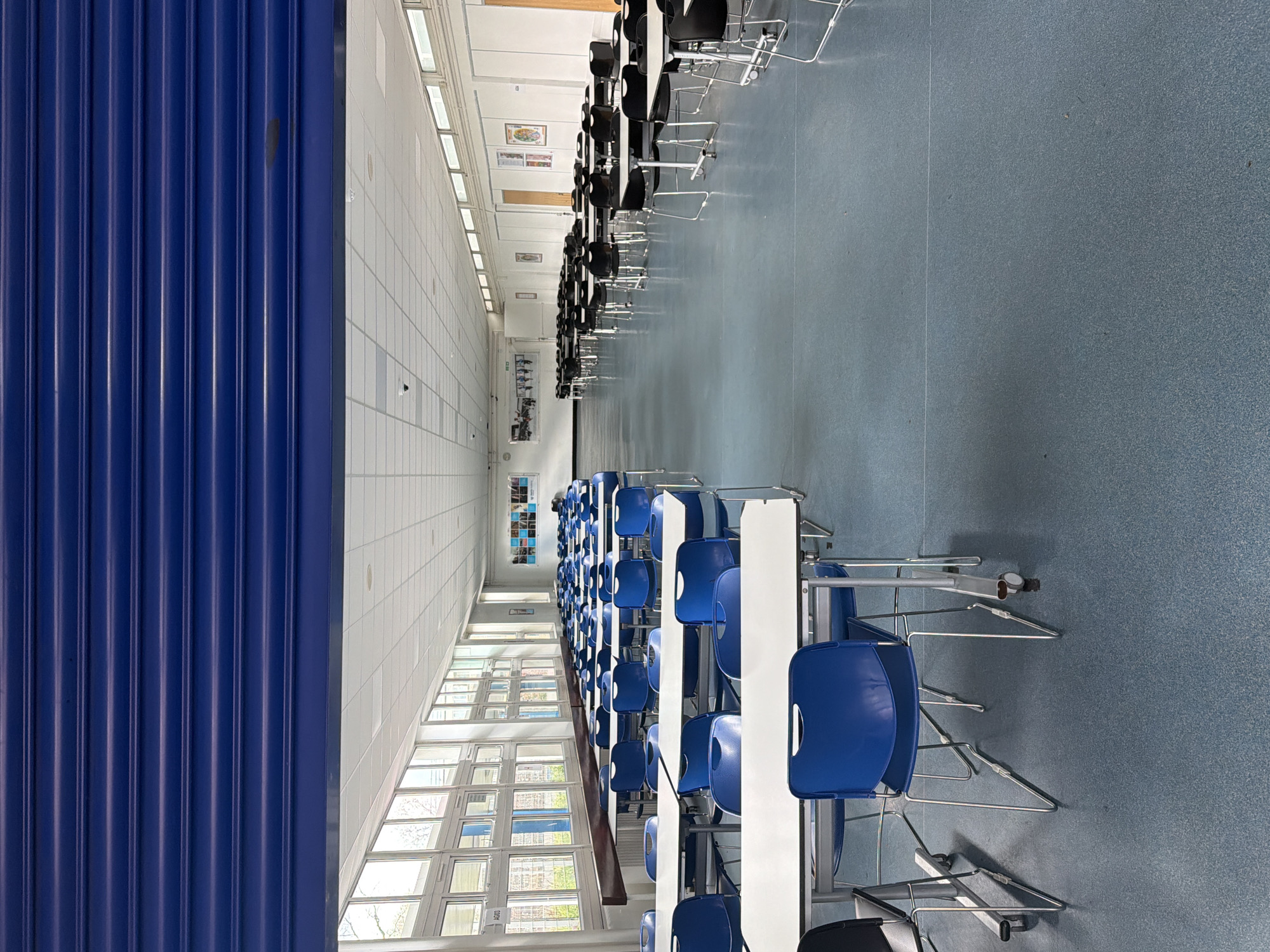 The image depicts an unoccupied institutional cafeteria or common room, specifically identified as likely located within the Macadam Building, King's College London, Strand Campus, London, United Kingdom.
The perspective is from an entrance, with a royal blue, horizontally slatted roller shutter partially lowered across the top of the frame. The floor is a uniform speckled blue-grey linoleum.
Along the left wall, a series of tall, rectangular windows with white frames extend above a dark brown wooden ledge. Through these windows, external views of bare tree branches and distant buildings are visible. A white label marked "AG03" is affixed to one of the window frames.
In the foreground and mid-ground on the left, multiple rectangular tables with white laminate tops and chrome-plated metal frames are arranged in rows. Each table is paired with several blue chairs, also featuring chrome-plated metal frames; some tables exhibit small caster wheels on their legs.
Towards the right and receding into the background, a separate section of identical white-topped rectangular tables is present, accompanied by black chairs, all constructed with chrome-plated metal frames. These are similarly arranged in rows.
The right wall is light-colored, featuring several closed wooden doors and various posters or framed graphic displays. The ceiling consists of a grid of white acoustic tiles, incorporating recessed rectangular light panels and multiple small, black dome-shaped security cameras. No individuals, actions, or interactions are present in the scene.