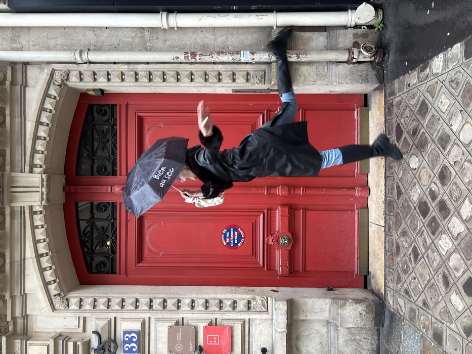 This image captures a person mid-jump in front of a distinct red double door at 33 Rue des Francs Bourgeois, in the 4th arrondissement of Paris, France. The subject, a woman viewed in profile, is depicted in dynamic motion, with her left foot lifted off the ground and her right leg bent upward behind her, suggesting a skip or leap. She wears a dark, possibly black, long coat with a prominent fur collar, light blue jeans, and black knee-high boots. In her raised left hand, she holds an open black umbrella featuring the white text "Bien au sec" (Well dry); her right arm is extended forward. Her mouth is open in an expression of mirth or exertion.
The background is dominated by a richly detailed Parisian building facade constructed from light-colored, cut stone blocks. The striking red double door, deeply recessed within an arched opening, serves as the central focal point. It features an ornate, gilded lion-head knocker at chest height and an arched transom window above, filled with intricate black wrought iron grilles. A circular red, white, and blue sign affixed to the left door panel explicitly states, "PRIÈRE DE NE PAS STATIONNER DEVANT CETTE PORTE" (Please do not park in front of this door). To the left of the door, on the stone facade, a blue rectangular plaque displays the white number "33." Below it, a vertical stack of smaller, rectangular plaques, some appearing to be brass, are visible, with the letters "E.A.C." discernible on the bottom plaque. To the right of the door, a light grey vertical pipe system runs against the building's stone wall, connecting to a red cylindrical fixture near the damp, grey cobblestone sidewalk, which is composed of geometrically patterned square and diamond pavers. The wet pavement reflects ambient light, indicating recent precipitation.