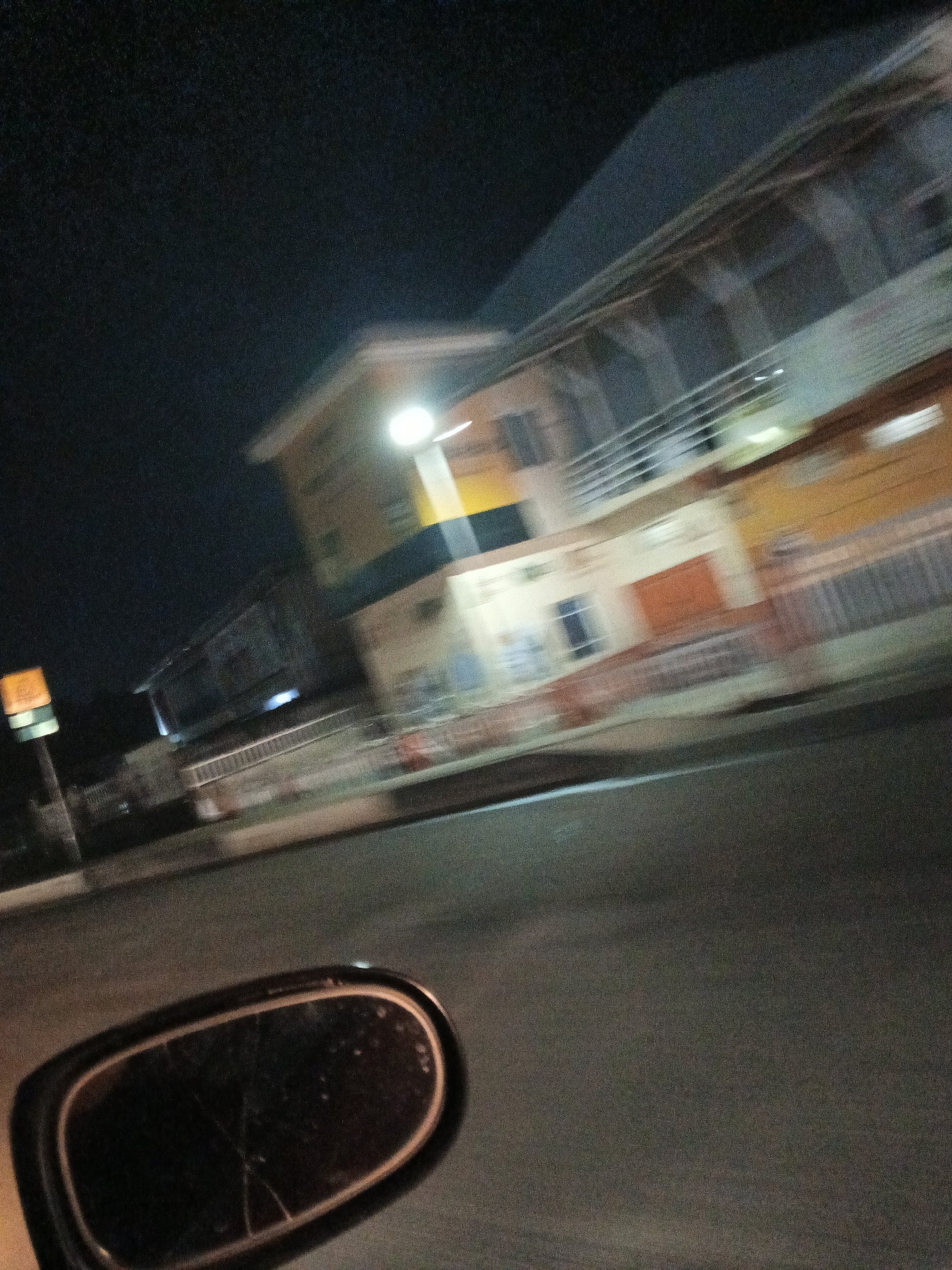 The image captures a nighttime scene in Jos, Nigeria, as suggested by the architecture and street lighting typical of the region. The photograph appears to have been taken from a moving vehicle, resulting in motion blur. In the foreground, a car's side mirror is visible on the left, reflecting a dark, indistinct scene. The midground features a two-story building with a light yellow and white facade, accented by a yellow and black horizontal stripe. The building has multiple windows and balconies with railings, and an orange door is visible. A street light illuminates the front of the building. In the background, more buildings and street elements are blurred due to motion. The overall environment is dark, indicative of nighttime, with artificial lighting from buildings and potentially streetlights casting a glow. The scene suggests a residential or commercial area in the city.