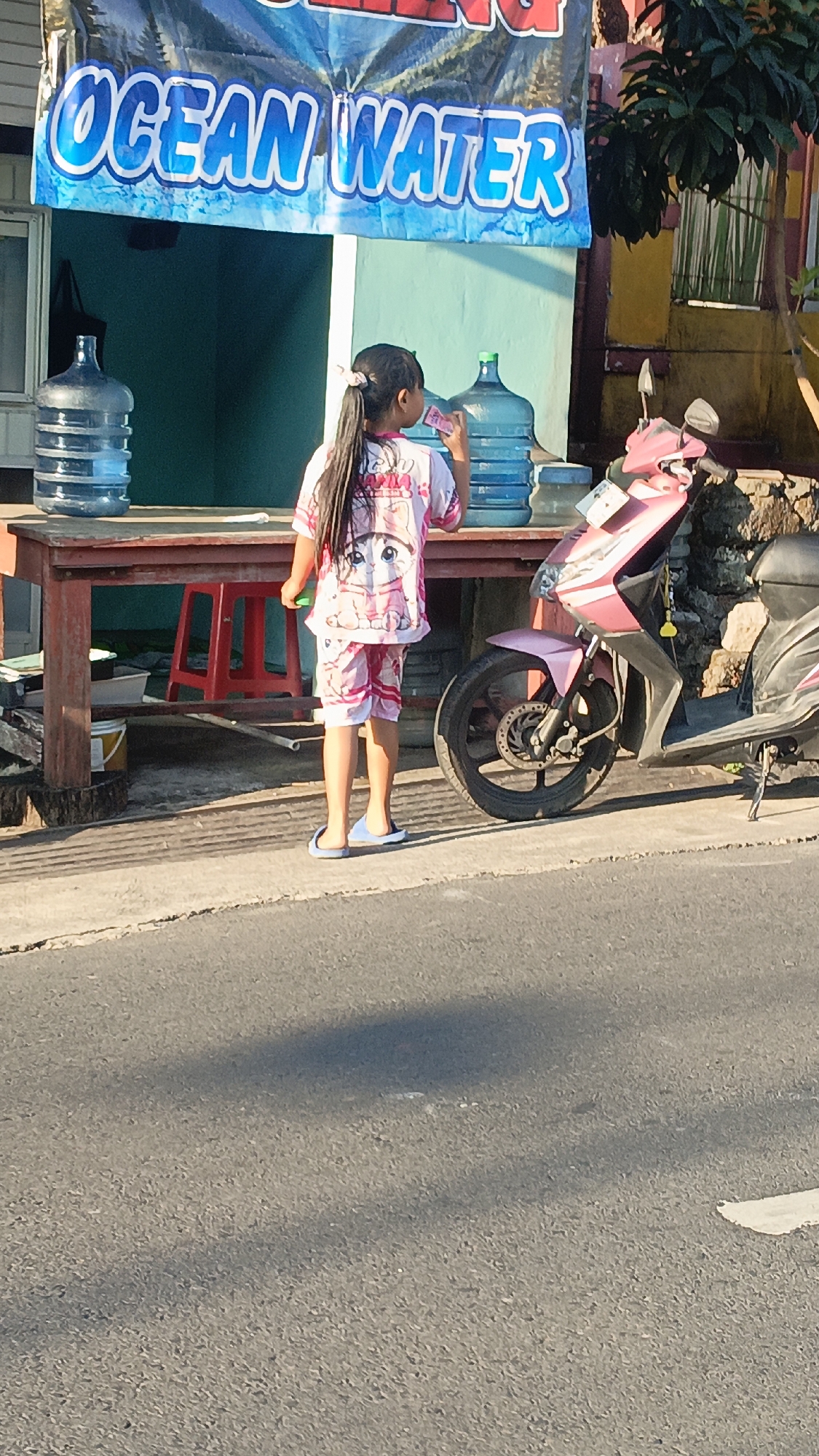A young girl with long, dark hair tied in a ponytail stands facing away from the camera, wearing a pink and white pajama set with a cat design. She is holding a small pink object and appears to be drinking from a cup. The girl is standing on a concrete sidewalk next to a red wooden table laden with large blue water dispenser jugs. A pink and black scooter is parked to her right. Behind the table, a blue banner with white lettering that reads "OCEAN WATER" is visible. The scene is set on a street, with asphalt in the foreground. The background includes a building with green and yellow sections and a tree. The image appears to have been taken in Salatiga, Indonesia, based on the context provided.