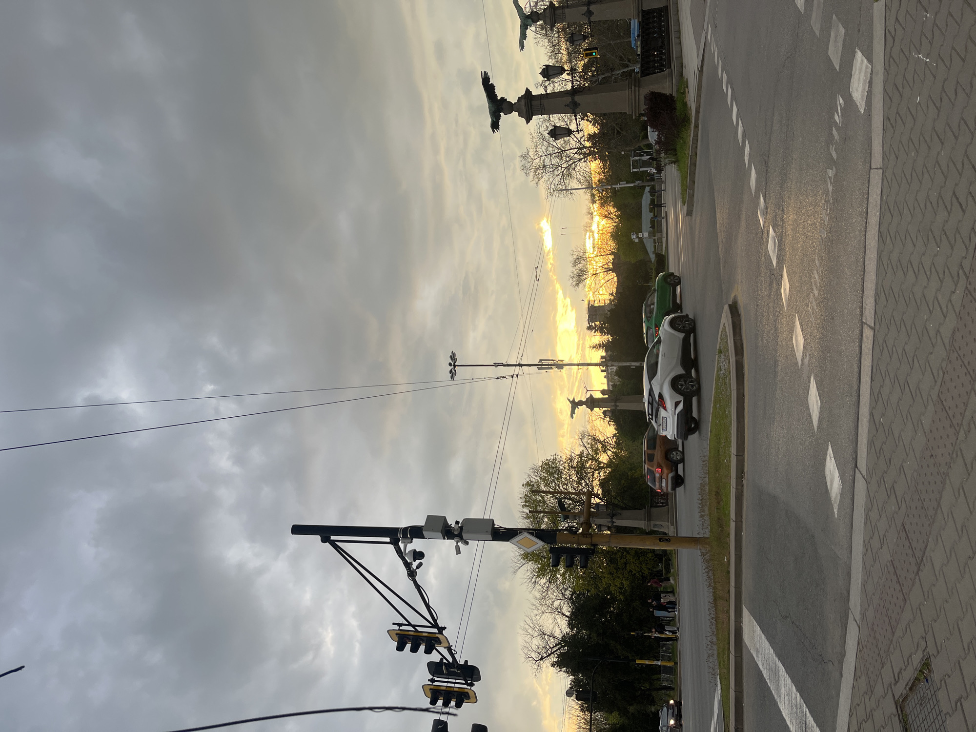 This landscape-oriented image depicts an urban street scene in Sofia, Bulgaria, likely captured during sunset or sunrise. The sky is largely overcast with extensive grey clouds, but a vibrant golden-yellow glow from the low sun breaks through the clouds on the horizon, illuminating the distant background and casting a warm hue over the scene.
In the foreground, a tall black pole dominates the left side, equipped with multiple traffic lights, some displaying a yellow signal, and several surveillance cameras. Overhead power lines stretch across the upper part of the frame.
The midground features an asphalt road with white dashed and solid lane markings, alongside a patterned grey paved sidewalk on the right. Several vehicles are visible: a white SUV and a green car are stopped or parked on the right side of the road, with a brown car further back. Trees, some with bare branches, line the street, suggesting either early spring or late autumn.
Distinctive monumental structures are silhouetted against the bright sky in the distance. These appear to be tall columns or pedestals topped with sculptures resembling large eagles or similar figures, characteristic of the Monument to Vasil Levski in Sofia. A few indistinct figures of people can be seen near these monuments.
The image conveys a sense of a quiet urban moment, with the dramatic natural light contrasting against the city infrastructure. No visible text is legible in the photograph.