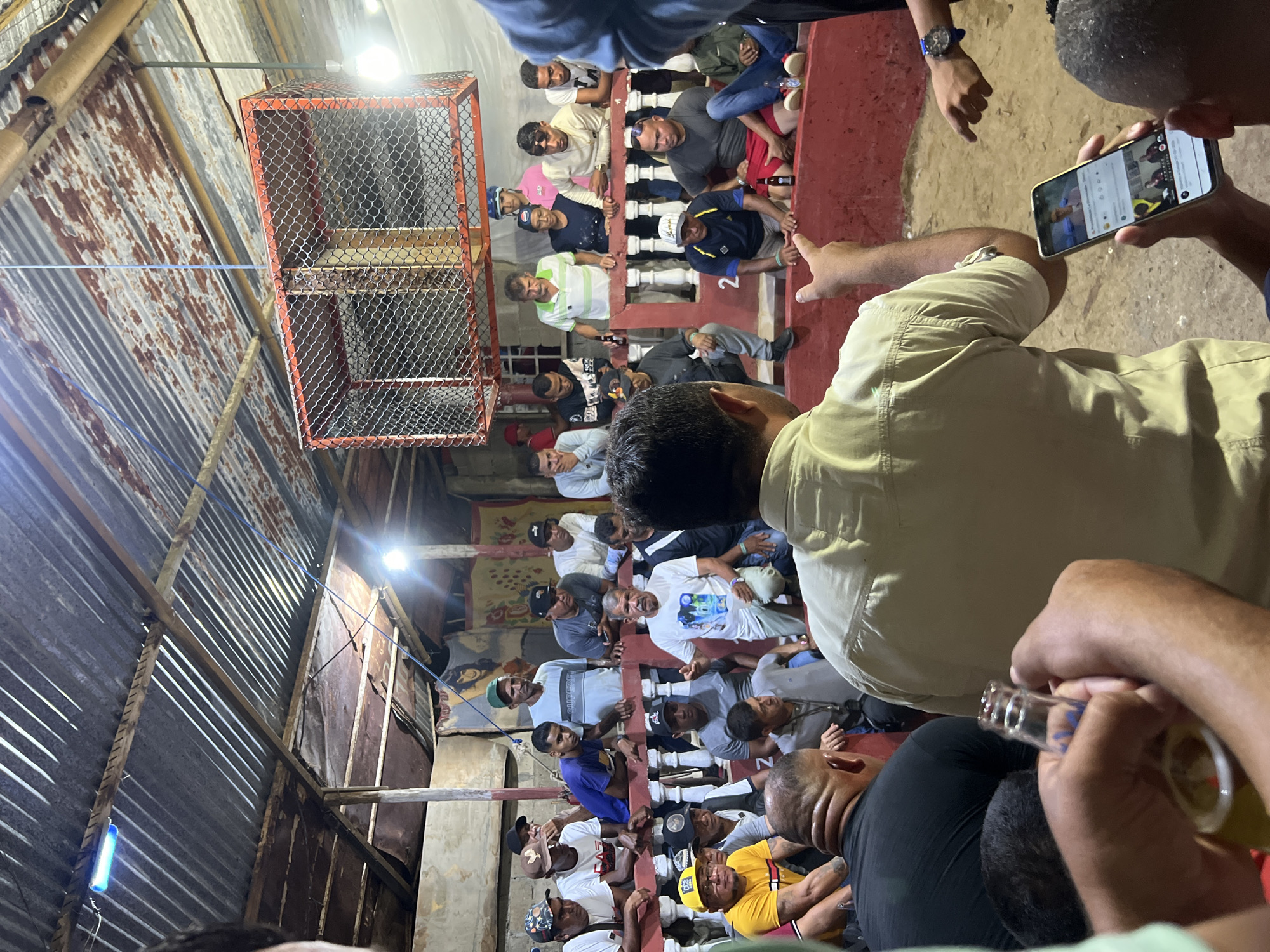 In Juan Griego, Venezuela, a gathering of men is observed in an outdoor, rustic arena with a corrugated metal roof. The participants are positioned around a central circular area, some leaning on a red railing. A cage-like structure, possibly for birds or animals, hangs suspended from the ceiling. The scene is illuminated by artificial lights. One individual in the foreground is holding a smartphone, displaying images and text. Another person is holding a bottle of beer. The atmosphere suggests a communal event or gathering.
