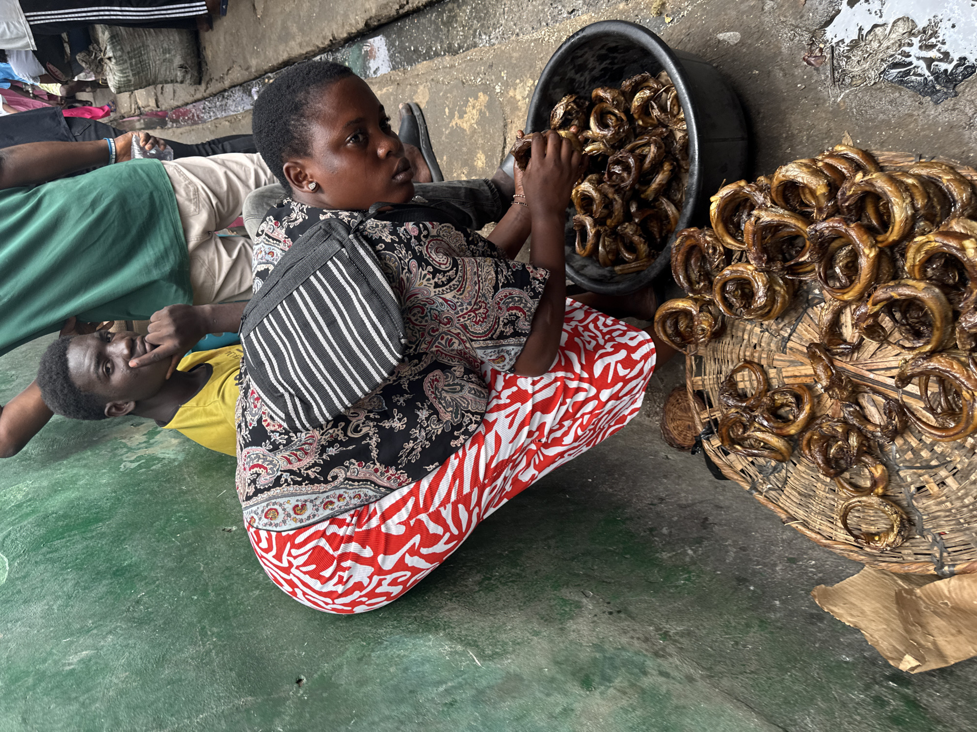 In a Lagos, Nigeria street scene, a woman is bent over, sorting through a large black basin filled with dried, coiled fish. She is wearing a black, patterned top with a black and white striped bag slung over her shoulder, and a vibrant red and white patterned skirt. To her left, a young man in a yellow t-shirt sits against a green, textured wall, looking towards the viewer with his hand near his chin. In the background, other individuals are visible, some standing, with a large sack and a drain visible on the paved ground. A woven mat, also piled with dried fish, lies on the ground in front of the woman. The overall environment suggests a bustling market or street-side vendor setting.