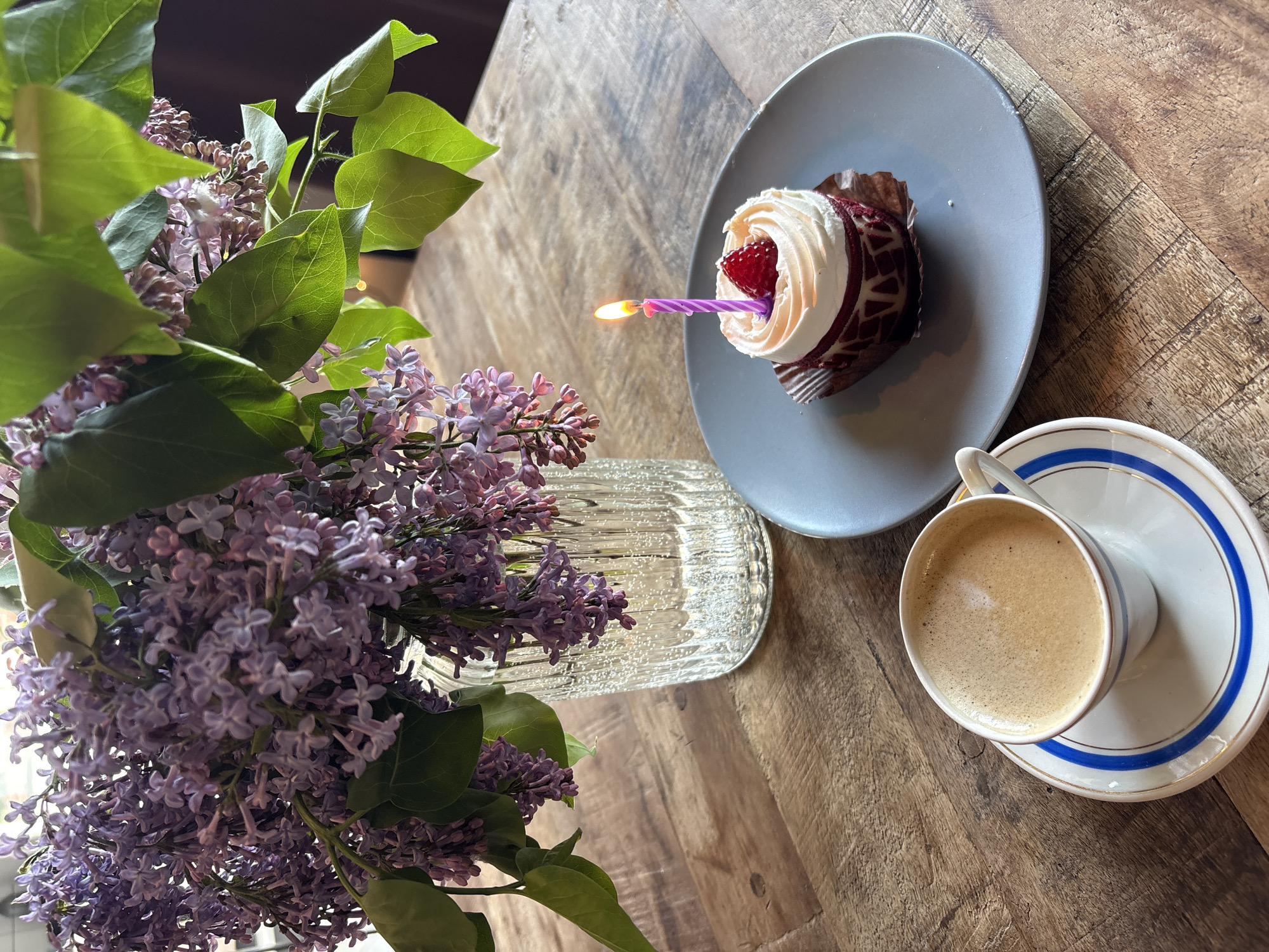 A high-angle shot captures a still life on a wooden table. A bouquet of lilac flowers in a clear, textured vase dominates the left side of the frame. To the right, a cupcake with white frosting and a red strawberry sits on a gray plate with a lit purple candle. Further to the right, a cup of coffee rests on a saucer with blue and gold trim. The scene is likely indoors, given the controlled lighting and composition, but the specific location in Saint Petersburg, Russia cannot be determined from the image alone. No people are visible.