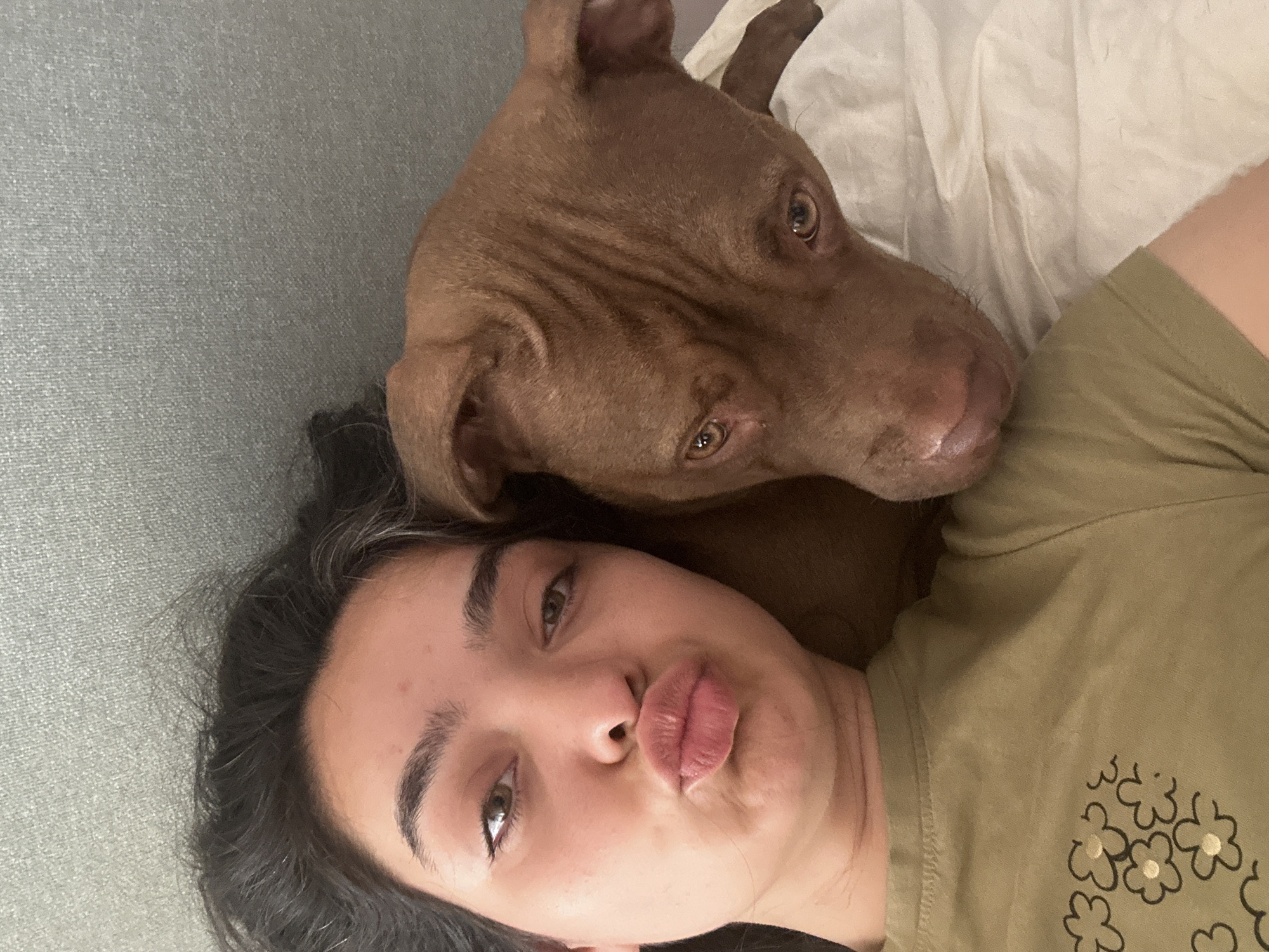 A close-up, overhead selfie shows a young woman lying down with her head on a pillow. She has dark hair and is wearing a green t-shirt with a floral design. Next to her, a brown dog rests its head on her shoulder, looking towards the camera. The background appears to be a gray textured surface, possibly a headboard or wall, and white bedding. The scene is indoors, with the soft lighting suggesting it might be a bedroom. There is no indication of the location being in the city of Malevo, Bulgaria.