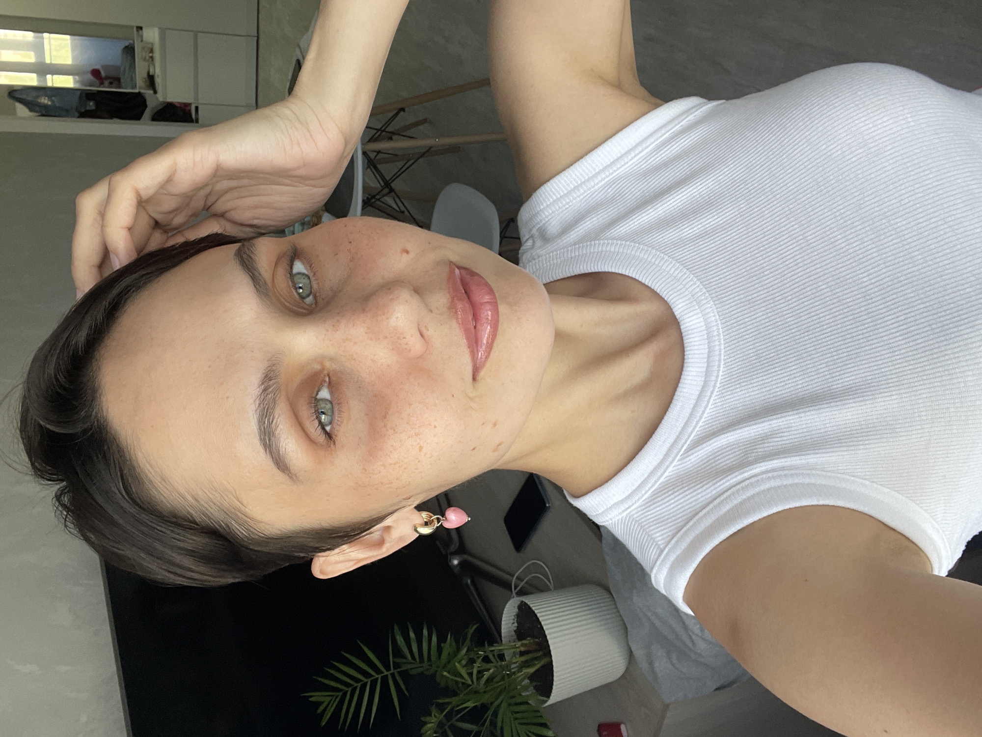 A woman with fair skin, freckles, and short dark hair takes a selfie. She is wearing a white ribbed tank top and pink heart earrings. Her hand is near her head, and she is looking at the camera. In the background, there is a room with light-colored walls, a white chair, and a potted plant. A black object, possibly a phone, is on a surface. There's a view of a building through a window, suggesting an indoor location. It is not possible to determine if the photo was taken in Izhevsk, Russia.