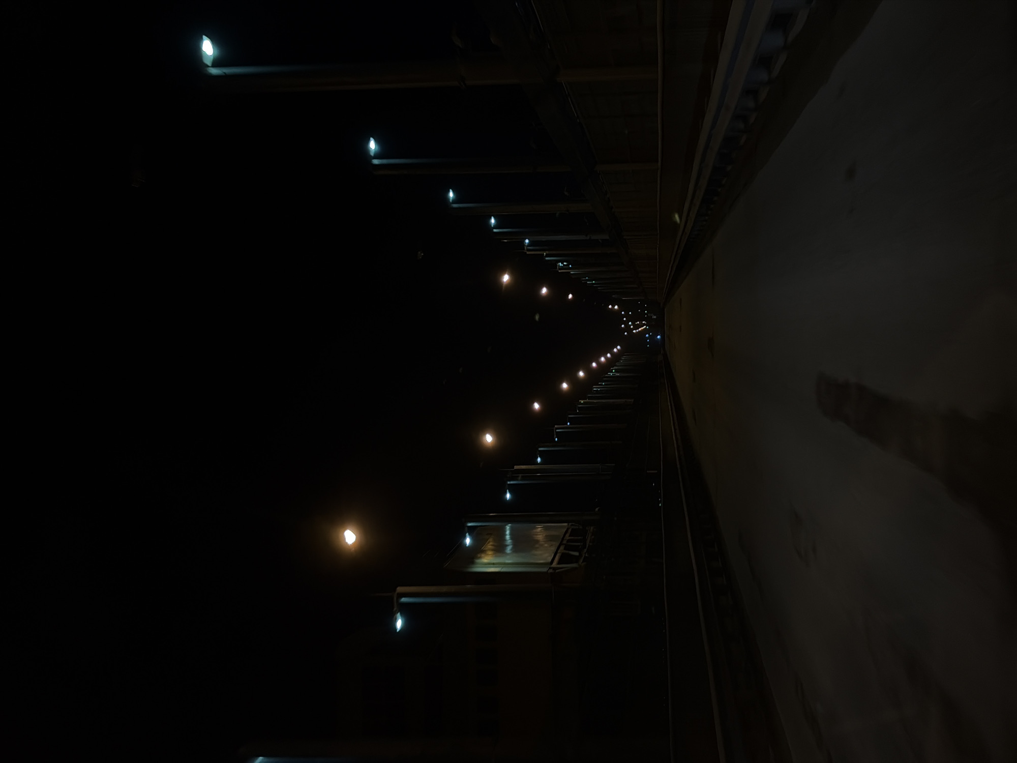 A low-angle shot in Chaikovsky, Russia, possibly a service tunnel or underpass, shows a dark corridor stretching away into the distance. The left side of the image is dominated by darkness, while the right is a wall that is lightly lit. A series of regularly spaced lights illuminate the top of the left wall and the dark floor of the tunnel. The perspective emphasizes depth, with the lights diminishing into a vanishing point far down the corridor. The tunnel's construction appears to be utilitarian, with exposed beams and concrete.