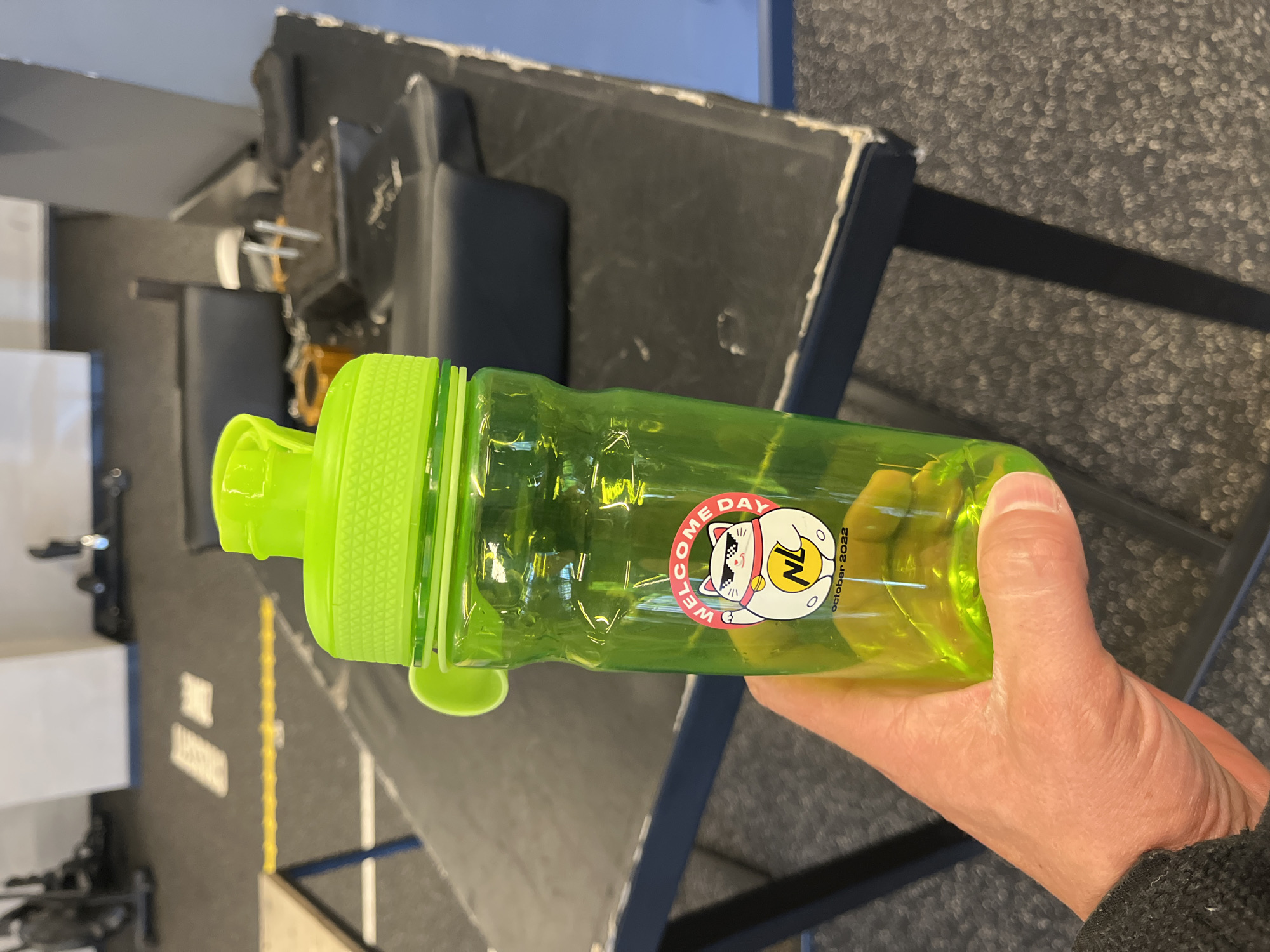 A close-up, eye-level shot in a fitness facility located in Sochi, Russia, captures a left hand holding a translucent lime green water bottle. The bottle features a bright green screw-on cap with a flip-top drinking spout and a small integrated carrying loop. Affixed to the front of the bottle is a circular sticker depicting a white Maneki-neko (waving cat) wearing black sunglasses. The sticker text around the top curve reads "WELCOME DAY" in red capital letters. On the cat's belly, the yellow letters "NL" are visible, and below the cat, "october 2022" is printed in black. The hand, visible from the wrist (covered by a dark grey sleeve) to the fingertips, grips the bottle. The background is out of focus, revealing a dark grey textured floor, possibly rubber gym matting, with a distinct yellow linear marking. Behind the floor, a dark, worn table or bench is visible, along with blurred shapes of exercise equipment and structures against light-colored walls.