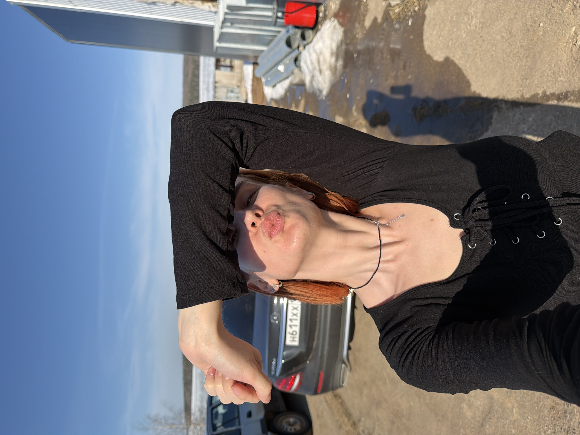 A woman is taking a selfie outdoors, likely in Chaikovsky, Russia, given the license plate on the dark-colored car behind her reads "H611XX." She is wearing a long-sleeved black top with a laced front. Her hair is reddish-brown. The background includes a building under construction with metal beams and pipes and a blue sky. Patches of snow and sunlight are visible on the ground.