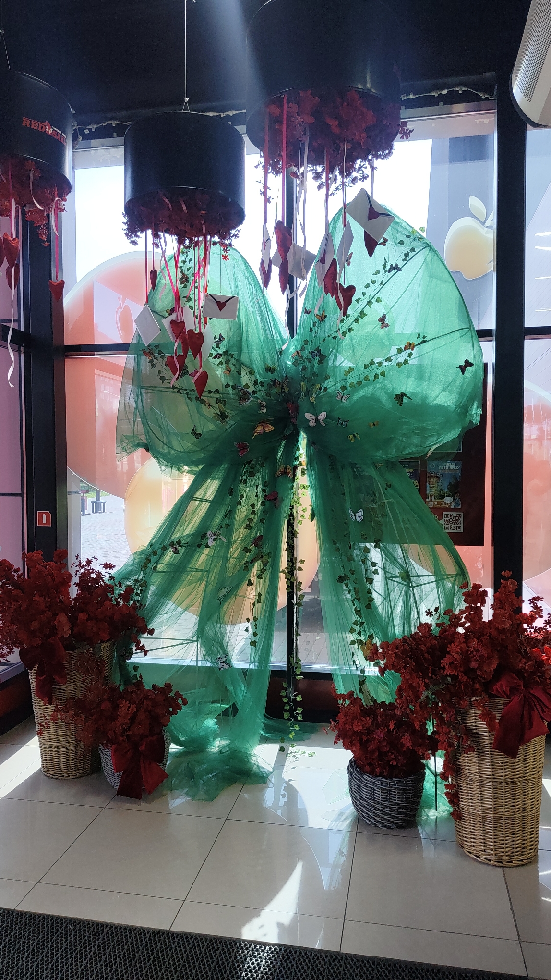 The image depicts an indoor decorative display, possibly for a special occasion or event. The central element is a large, voluminous green tulle bow, adorned with artificial ivy vines and numerous small butterflies. Hanging from above are dark cylindrical structures from which red ribbons and small red hearts, along with miniature white envelopes, descend. In the foreground, on either side of the bow, are large woven baskets filled with vibrant red floral arrangements. The floor is tiled, and sunlight streams in from large windows in the background, creating bright reflections. The windows reveal a glimpse of the exterior, including some large, out-of-focus orange spheres and a faint image of an apple logo. There is a red pull handle with a white circle on the left side of the frame, possibly a fire alarm or emergency door release. The scene appears to be located in a public or commercial space, such as a store or event venue. No specific information about Krasnoyarsk, Russia, is evident in the image.