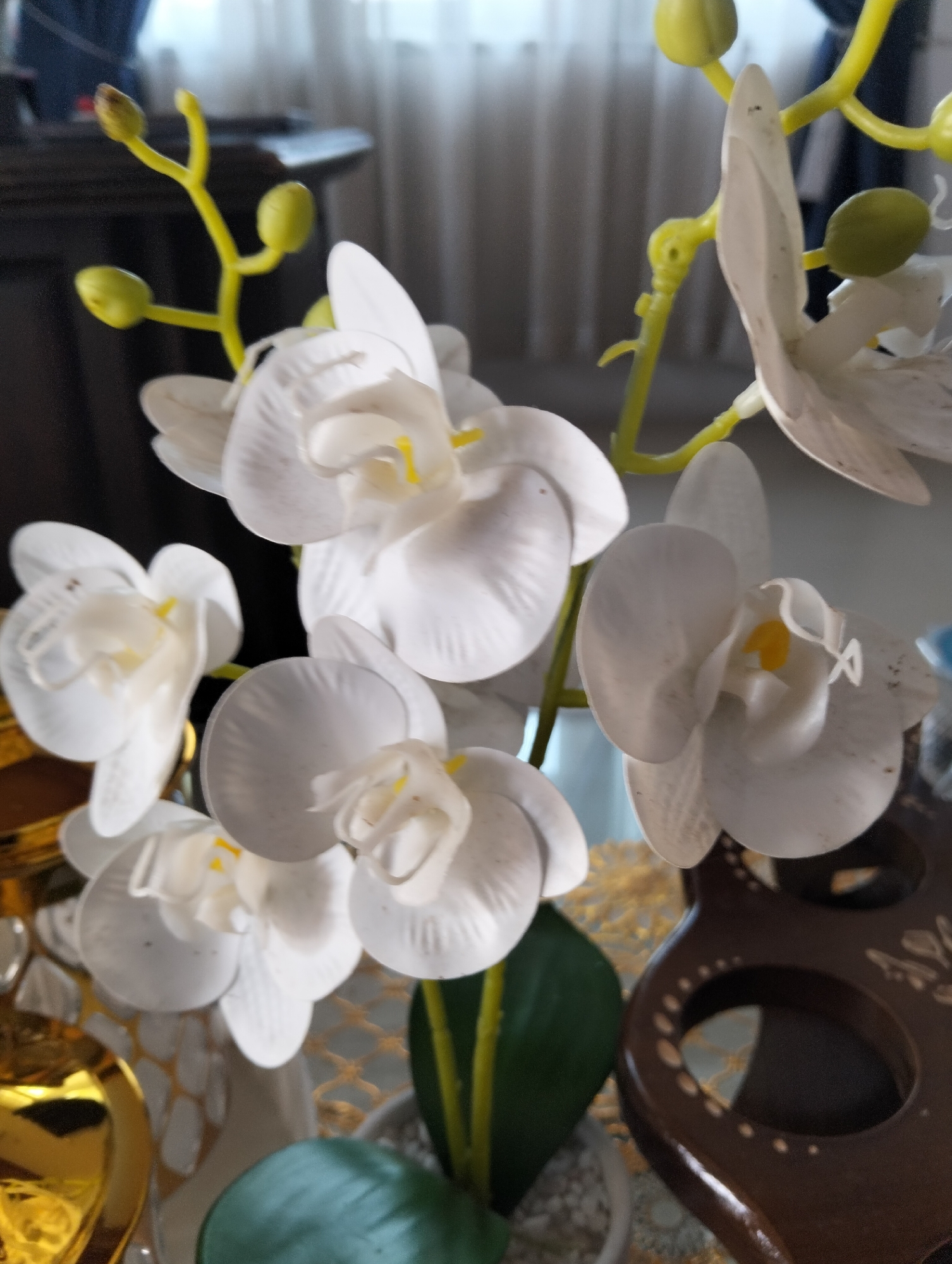 This image displays a close-up of several artificial white orchids with yellow centers, arranged on a stem with green buds. The orchids are in focus, with some appearing fully bloomed and others partially open. The background is blurred, showing a light-colored curtain and dark furniture. The scene appears to be indoors, likely on a table or shelf, given the presence of decorative objects like a gold-colored vase or accessory on the left and a dark brown, carved wooden object with circular cutouts on the right. The overall impression is of a still life composition, possibly a home or office decor arrangement. The location in Julicotmeurak, Indonesia is not discernible from the visual elements of the image.