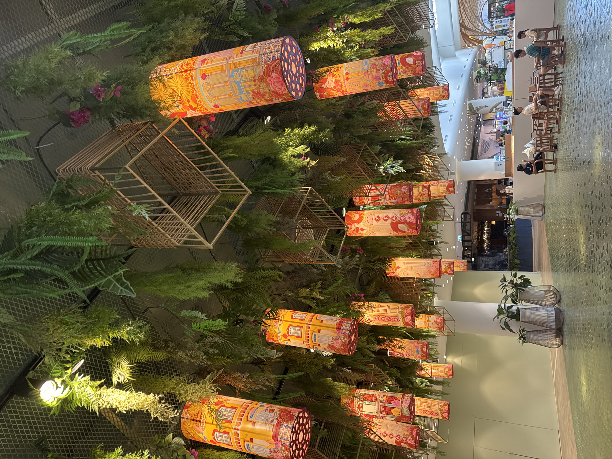 This image depicts the interior of a modern shopping mall, specifically in Wichit, Thailand. The ceiling is heavily adorned with lush green hanging plants and numerous cylindrical lanterns. These lanterns are predominantly yellow and feature intricate designs that appear to be depictions of traditional architecture with windows and floral motifs. Scattered among the plants are also geometric, cage-like structures made of wood or bamboo, adding to the decorative element. In the foreground, a group of four individuals are seated at low wooden tables and chairs, appearing to be engaged in conversation or relaxing. The floor is covered in a mosaic-like pattern of green tiles. To the left, there are large decorative vases containing plants. The background reveals a brightly lit shopping environment with various storefronts and escalators, indicating a bustling commercial space. The overall ambiance is one of vibrant decoration and a relaxed social gathering within a commercial setting.