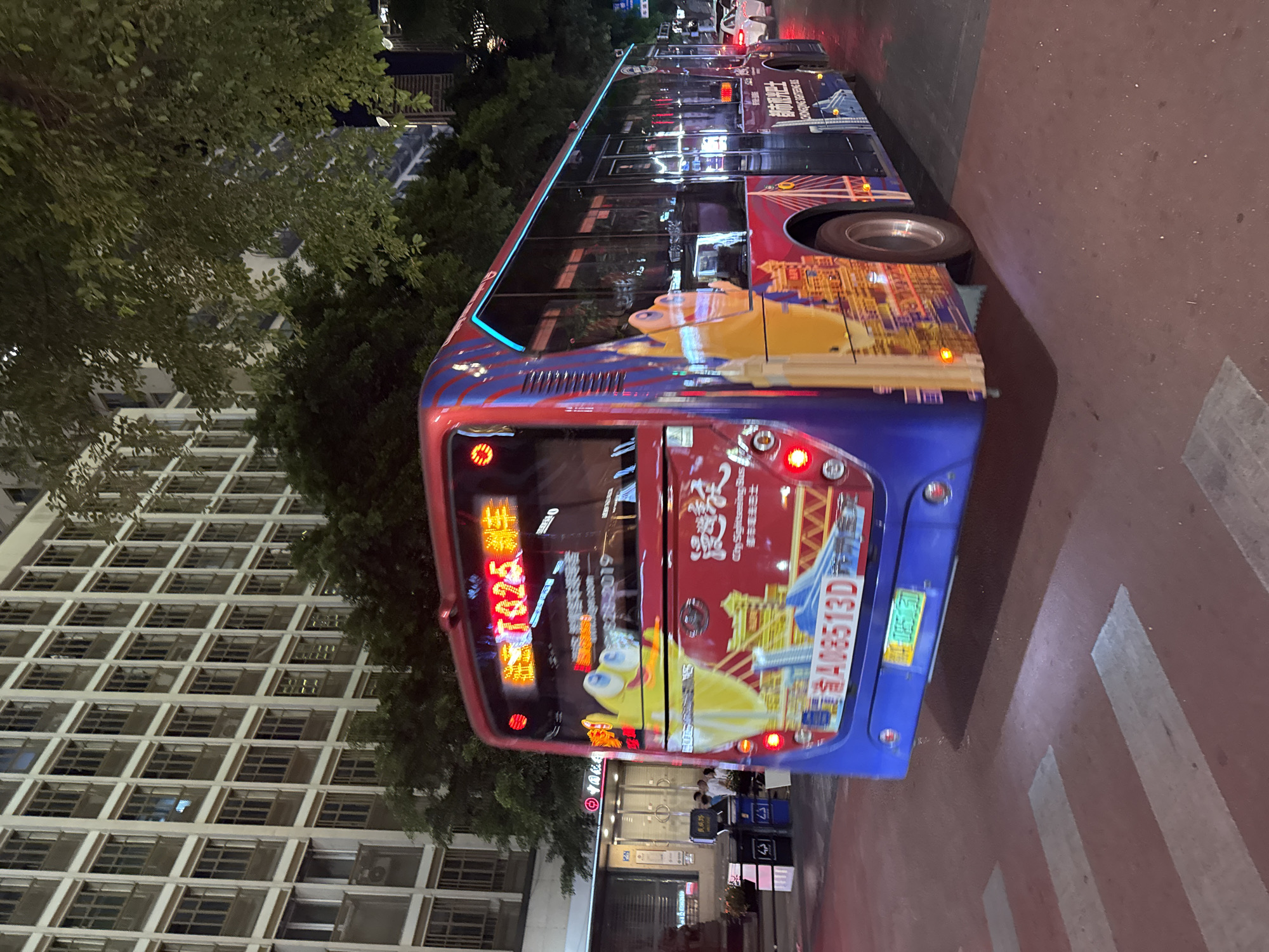 A blue and red bus, identified as a "Chongqing Sightseeing Bus" with the license plate "渝A08513D", is captured in motion on a city street at night. The bus features colorful artwork, including a cartoon character and cityscapes. It is illuminated by streetlights and the lights from a tall, multi-story building in the background. Trees with green foliage partially obscure the building. The bus is positioned on a crosswalk marked with red pavement and white stripes. In the background, a few individuals are visible near trash receptacles outside a storefront. The scene appears to be in Yuzhong District, China.
