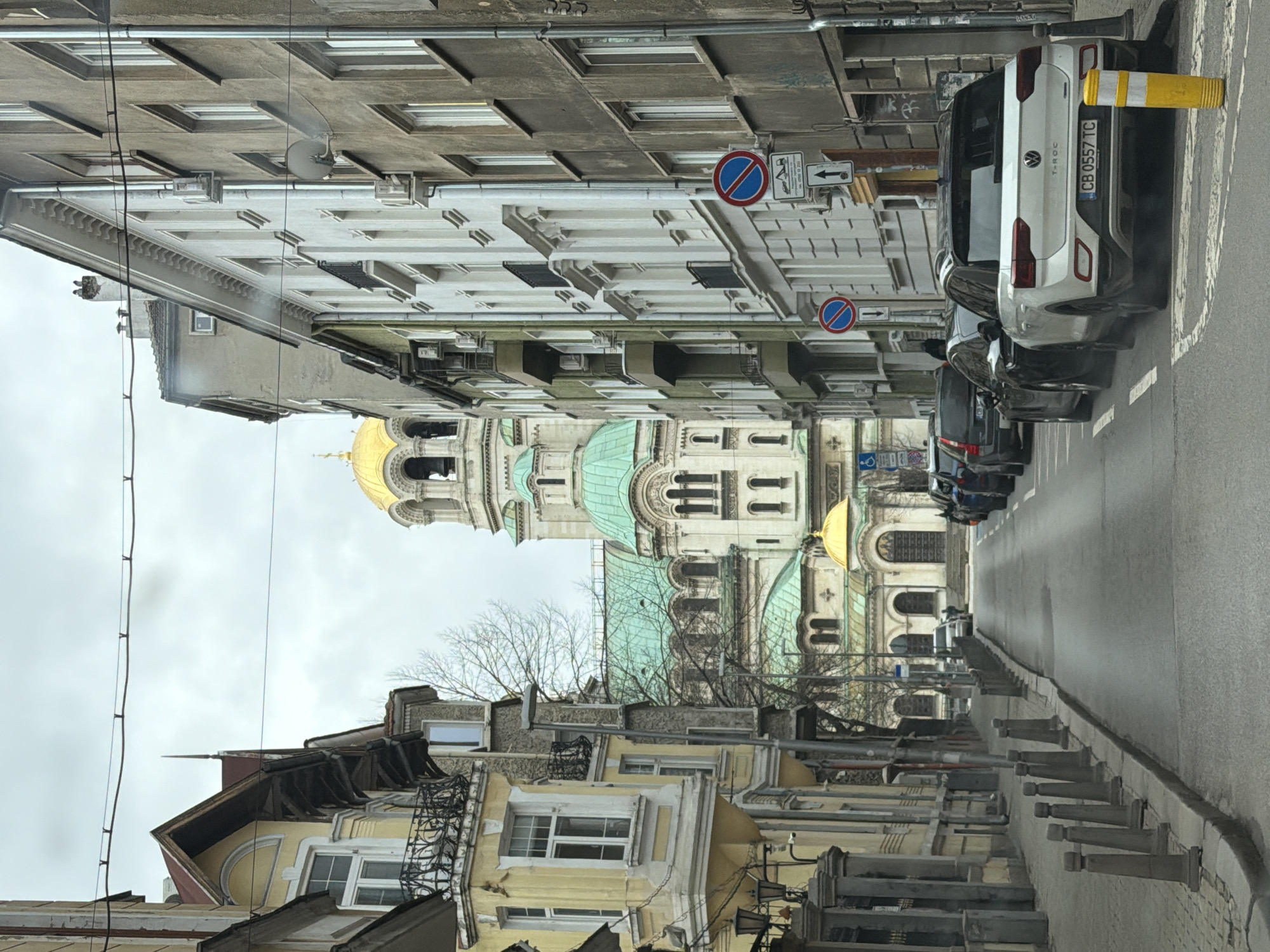 The image shows a street view in Sofia, Bulgaria, with the Alexander Nevsky Cathedral visible in the distance. The street is lined with buildings, and several parked cars are on the right side. A white VW T-ROC is parked closest to the viewer with the license plate number CB 0557 TC. Road signs, including a "no stopping" sign with a tow-away symbol and an arrow pointing to the left, are visible on the right. The sky is overcast.