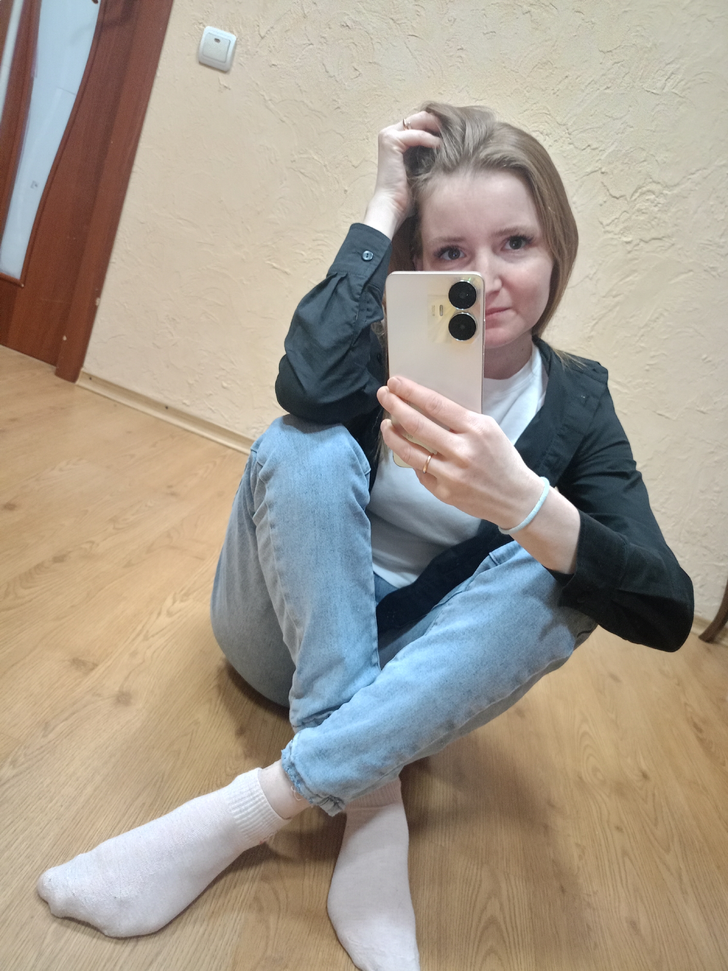 A light-skinned woman is sitting cross-legged on a light wood floor, holding up a smartphone in her right hand, as if taking a selfie. She wears light blue jeans, white socks, a white t-shirt, and a black button-down shirt, unbuttoned. Her hair is blonde and shoulder-length. Her left hand is in her hair. The background consists of a light-colored wall and a dark wooden door in the upper left corner.