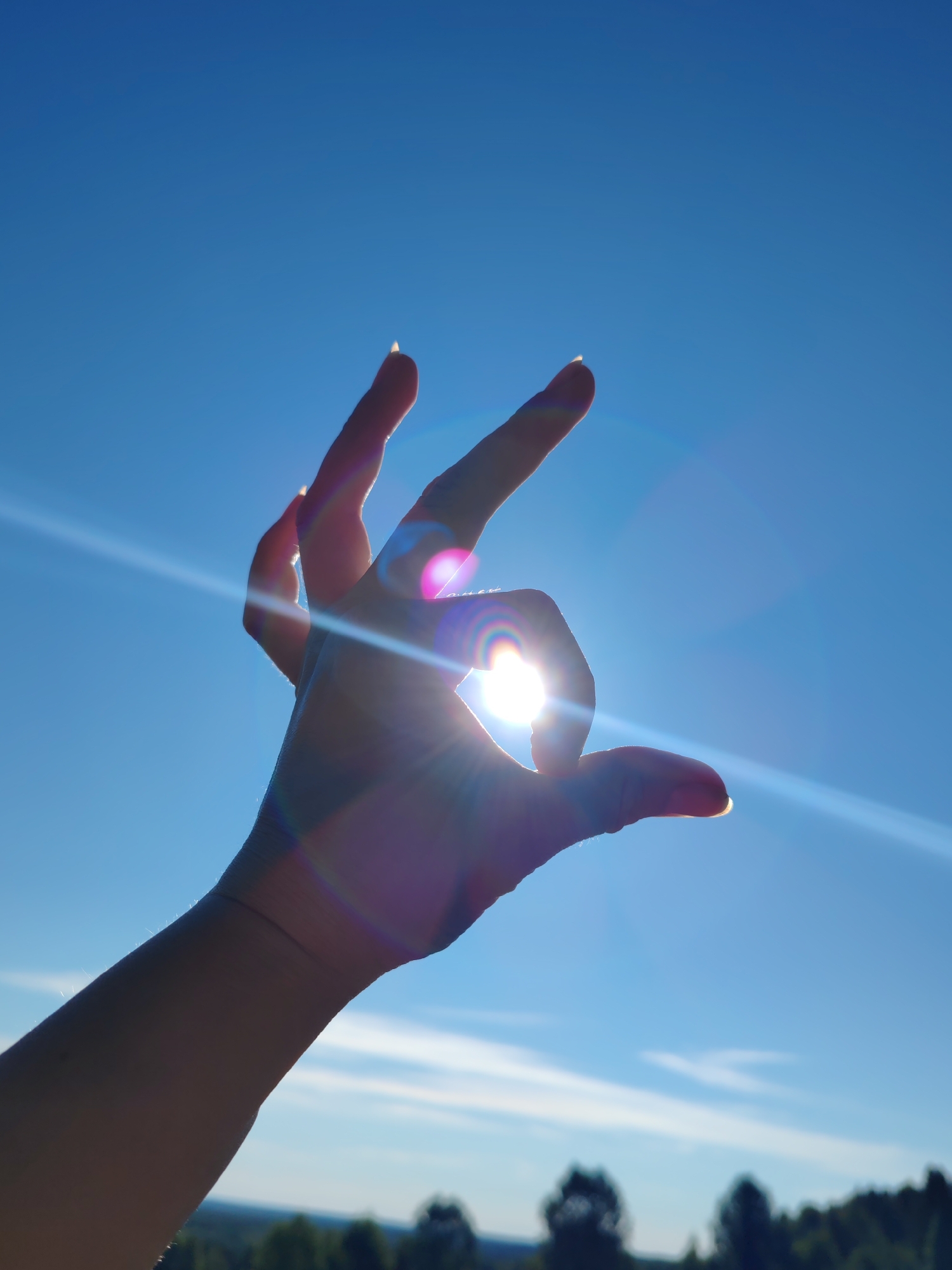 A human hand is silhouetted against a bright blue sky, with the sun shining through the gap between the thumb and forefinger, creating a lens flare effect. The hand is positioned in the foreground, with the fingers extended upwards and slightly curved. The background shows a blurry landscape of trees and a hint of clouds. The overall impression is one of a sunny day, possibly taken outdoors. This image does not provide any information about the location in the city of Чухлэм, Russia, or any specific urban context.