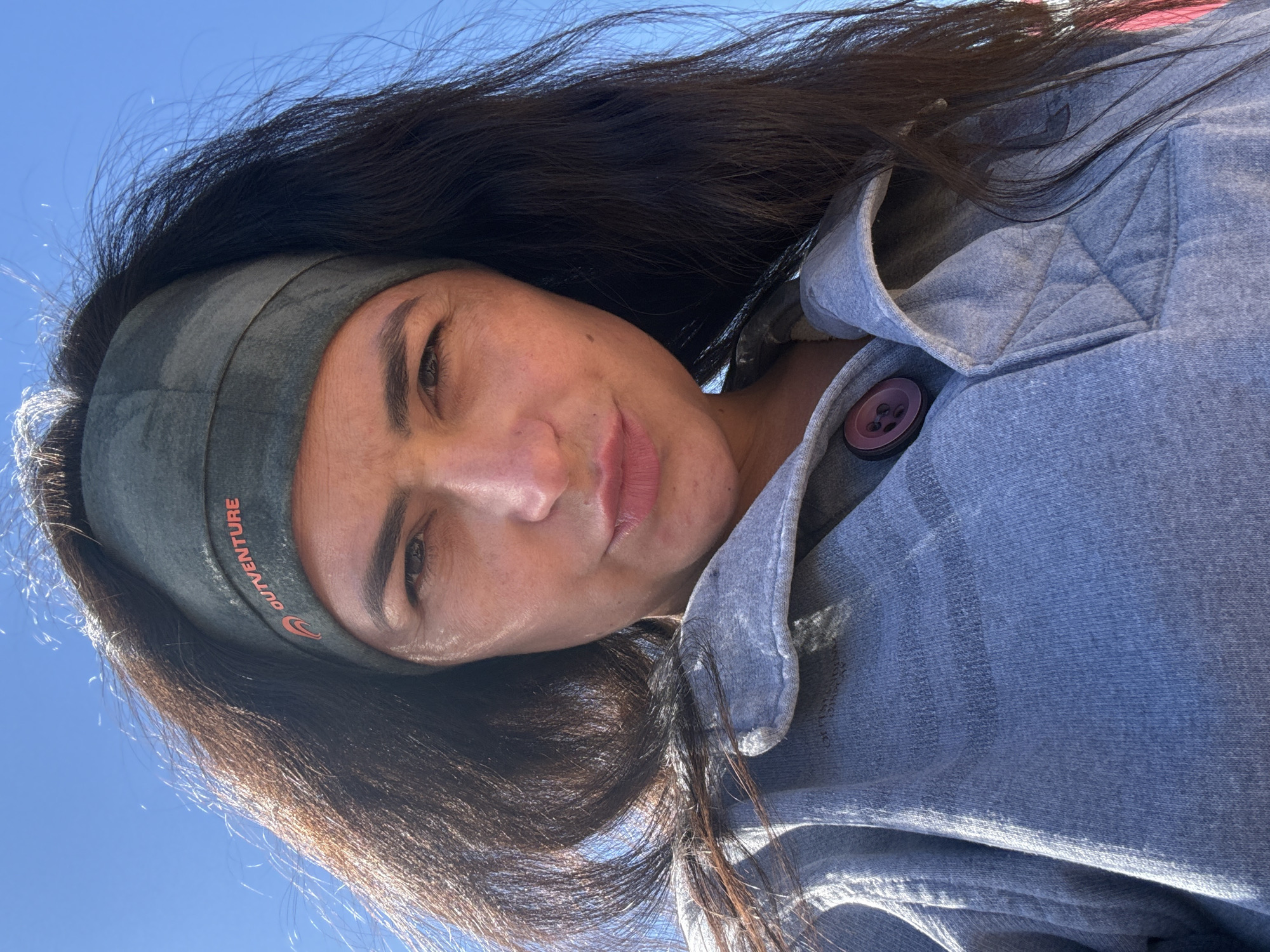 A woman with long brown hair wearing a gray headband with "OUTVENTURE" written on it, and a blue button-up jacket, takes a selfie against a bright blue sky. The location is not discernible.