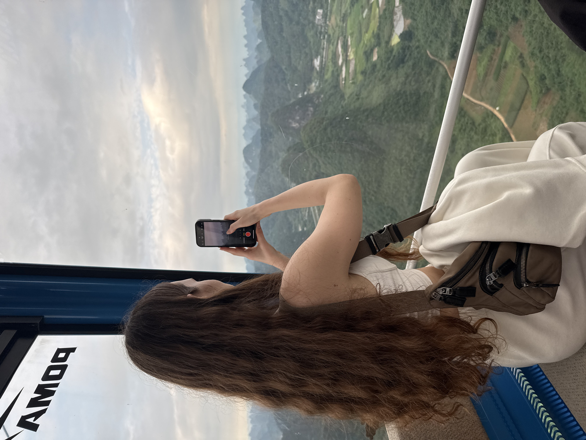 A woman with long brown hair is riding in a cable car in Guilin, China, recording a video with her phone. The landscape outside the cable car reveals a mountainous region with terraced fields. The woman is wearing a white top and a light-colored sweatshirt, with a small brown bag across her body. The inside of the cable car is blue and white, with the word "PONIA" visible on the interior.