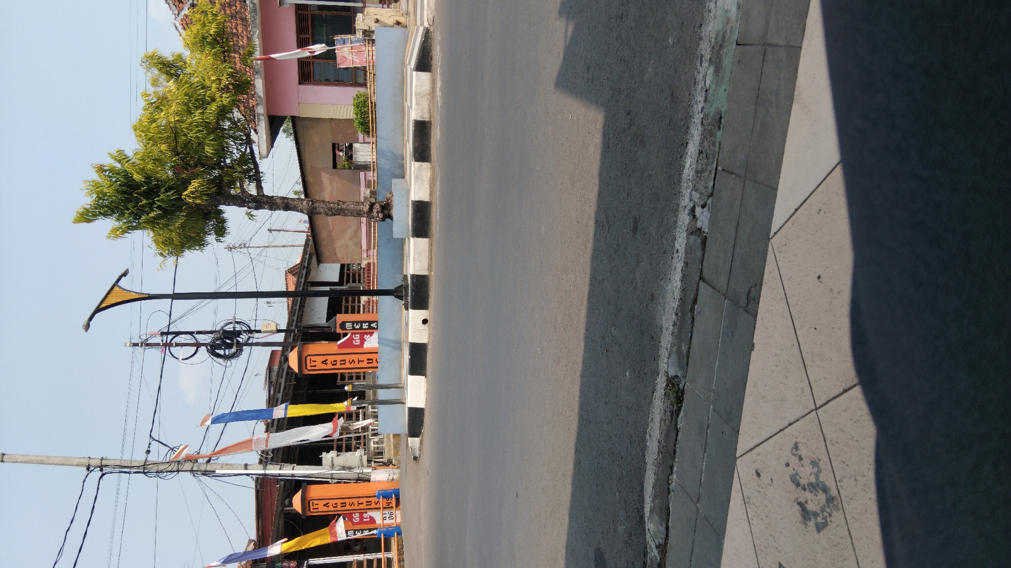 The image shows a street scene in the city of Batang, Indonesia, on a sunny day with a clear blue sky. The foreground is dominated by a paved road, with a sidewalk running along the right side of the image. The sidewalk is made of square tiles, some of which are cracked or discolored. A large shadow falls across the sidewalk and road, indicating the sun is high in the sky.
In the middle ground, there is a street curb with black and white stripes. Behind the curb, there are several orange pillars with Indonesian flags and text that appears to be related to the Indonesian Independence Day ("17 Agustus"). There are also several other flags in various colors. A modern-looking street lamp with a yellow shade stands near the pillars. A tree with green foliage grows near the street lamp. In the background, there are buildings, including a pink one with a tiled roof, and utility poles with many wires. The overall impression is of a typical street in a town or city in Indonesia.