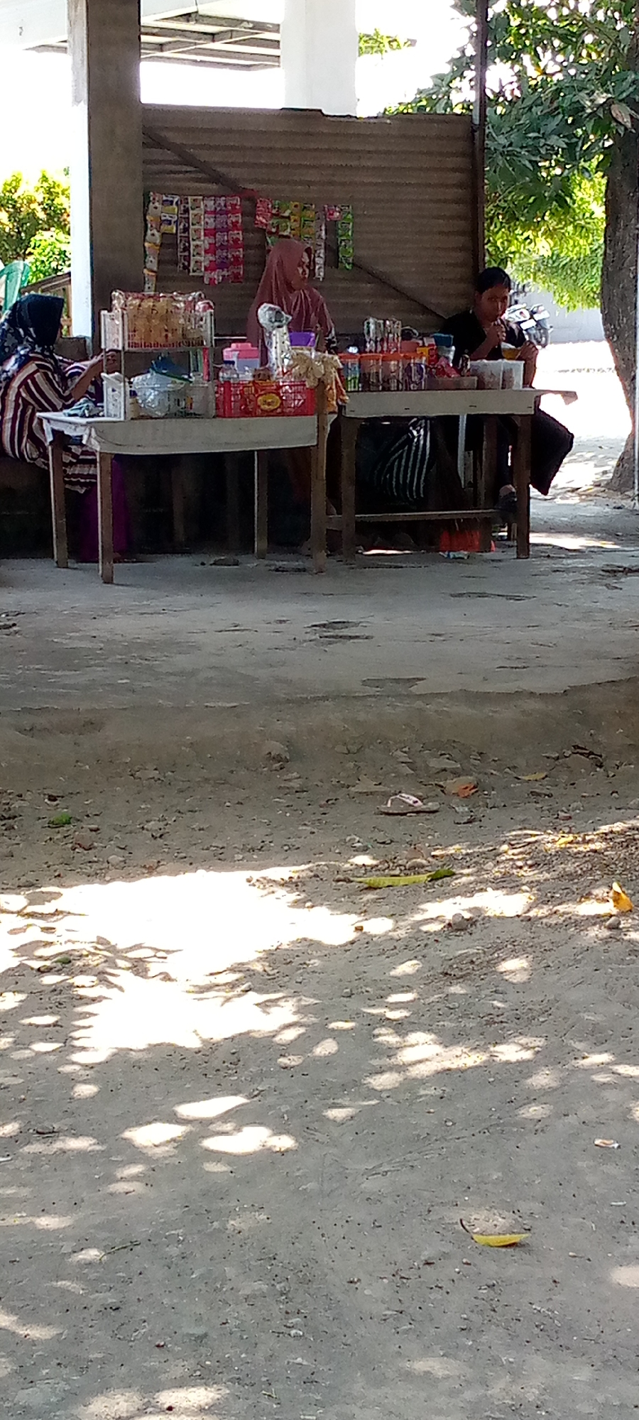 In Pacing, Indonesia, a street-side stall displays various packaged goods, including snacks and drinks. Two women are present at the stall, one wearing a pink hijab and the other a striped headscarf and dress. A young man, wearing a black t-shirt, sits on a stool at the stall, appearing to be eating or drinking. The stall is constructed of corrugated metal, and the ground in front of it is dirt. Sunlight casts dappled shadows from overhead trees onto the ground. A motorcycle is partially visible in the background.