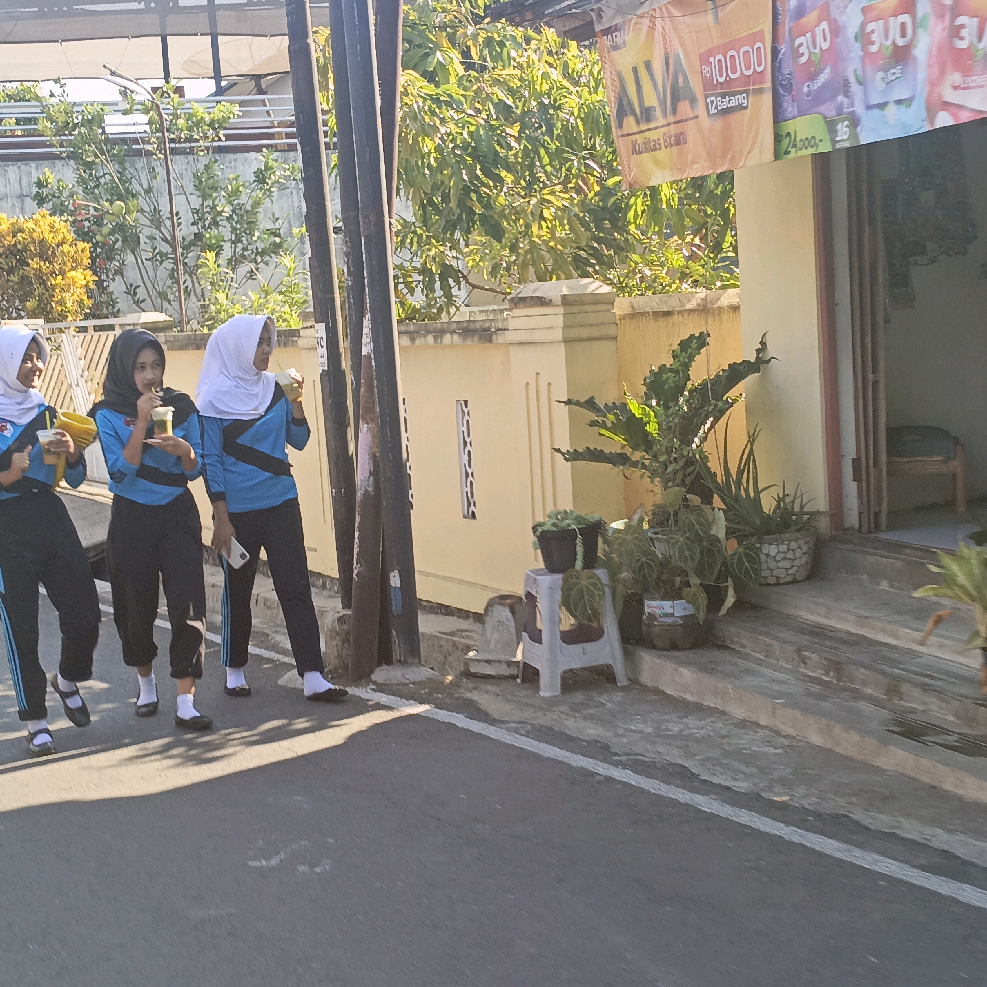 The image depicts a street scene in Salatiga, Indonesia, with three young women walking on the left side of the frame. They are wearing matching blue and black uniforms with white hijabs. Two of the women are holding and drinking from clear plastic cups. The woman on the far left is holding a yellow plastic container. They are walking on an asphalt road with a white painted line on the right side, separating the road from a sidewalk or paved area. In the background, there is a building with a yellow facade and a banner advertising "ALVA" with prices. Several potted plants are visible on a raised concrete area in front of the building. Sunlight casts shadows on the road, indicating it is daytime. The overall impression is a candid snapshot of daily life in a residential or commercial area.