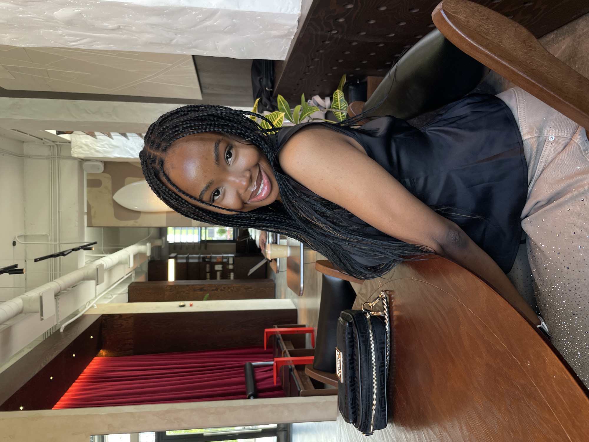 A young Black woman with braided hair smiles at the camera, seated at a wooden table in what appears to be a cafe. She is wearing a black sleeveless top and beige pants adorned with rhinestones. To her left on the table is a black clutch purse with a gold chain strap. The background shows a dimly lit interior with red curtains, chairs, and architectural elements. The overall scene suggests a casual, social setting in an urban environment.