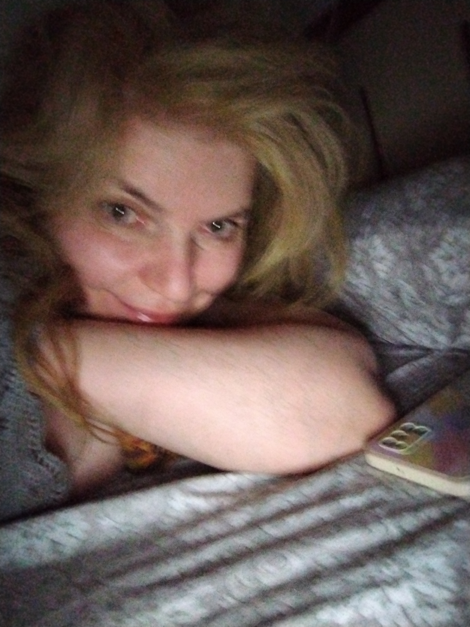 A person with blonde hair is lying on a bed. Their head is resting on their arm, which is placed on the bed. A mobile phone is near their arm on the bed. The bed has grey striped bedding. The environment appears to be indoors and dimly lit.
There is no evidence to suggest this image was taken in Al Mas'oudi, United Arab Emirates.