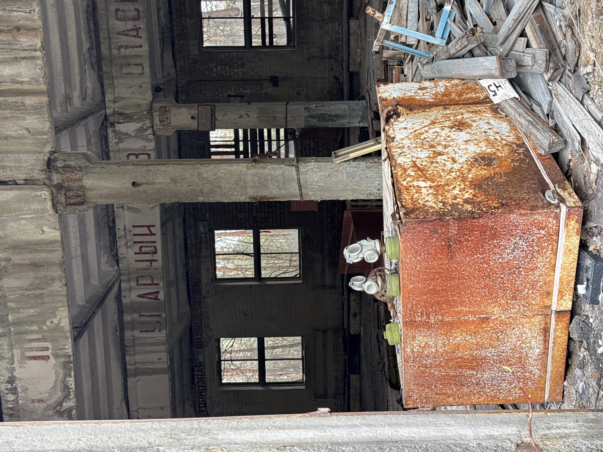 The image shows an interior of a derelict building in Chornobyl, Ukraine. Visible are exposed brick walls with several barred window openings. Concrete support pillars with fading red Cyrillic lettering run vertically along the image. A rusted metal box with attached gas masks rests in a pile of debris.