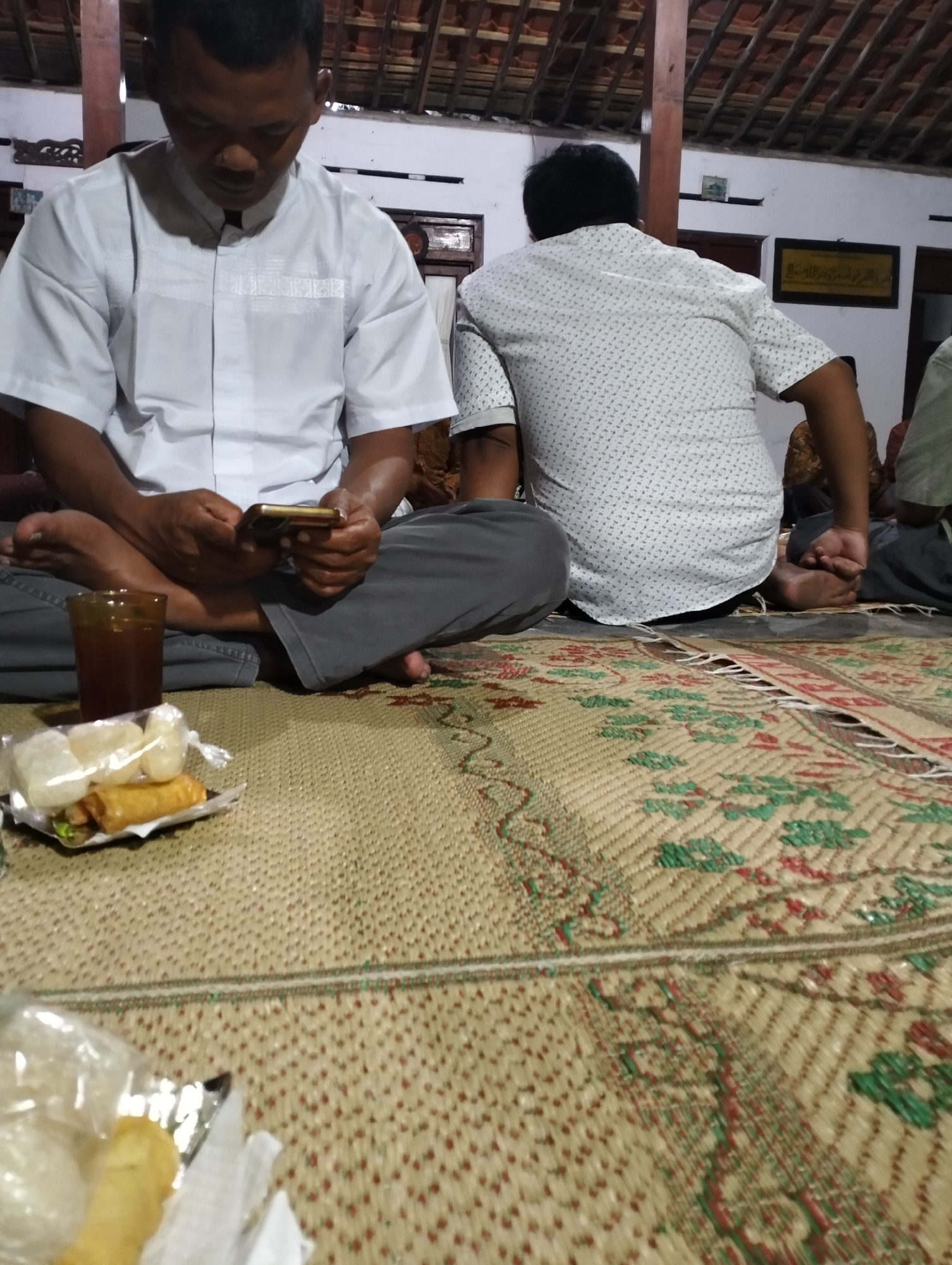 In Juwiring, Indonesia, two men are seated on a woven mat inside a building with a tiled roof. The man in the foreground, wearing a white shirt, is looking at his phone while holding it with both hands. In front of him is a glass of dark liquid and a small plate of food containing a spring roll and white, puffed snacks. To his right, another man is seen from the back, wearing a patterned shirt and also seated on the mat. The environment appears to be a room with white walls, and in the background, a framed artwork hangs on the wall. The mat has a decorative pattern of red and green.