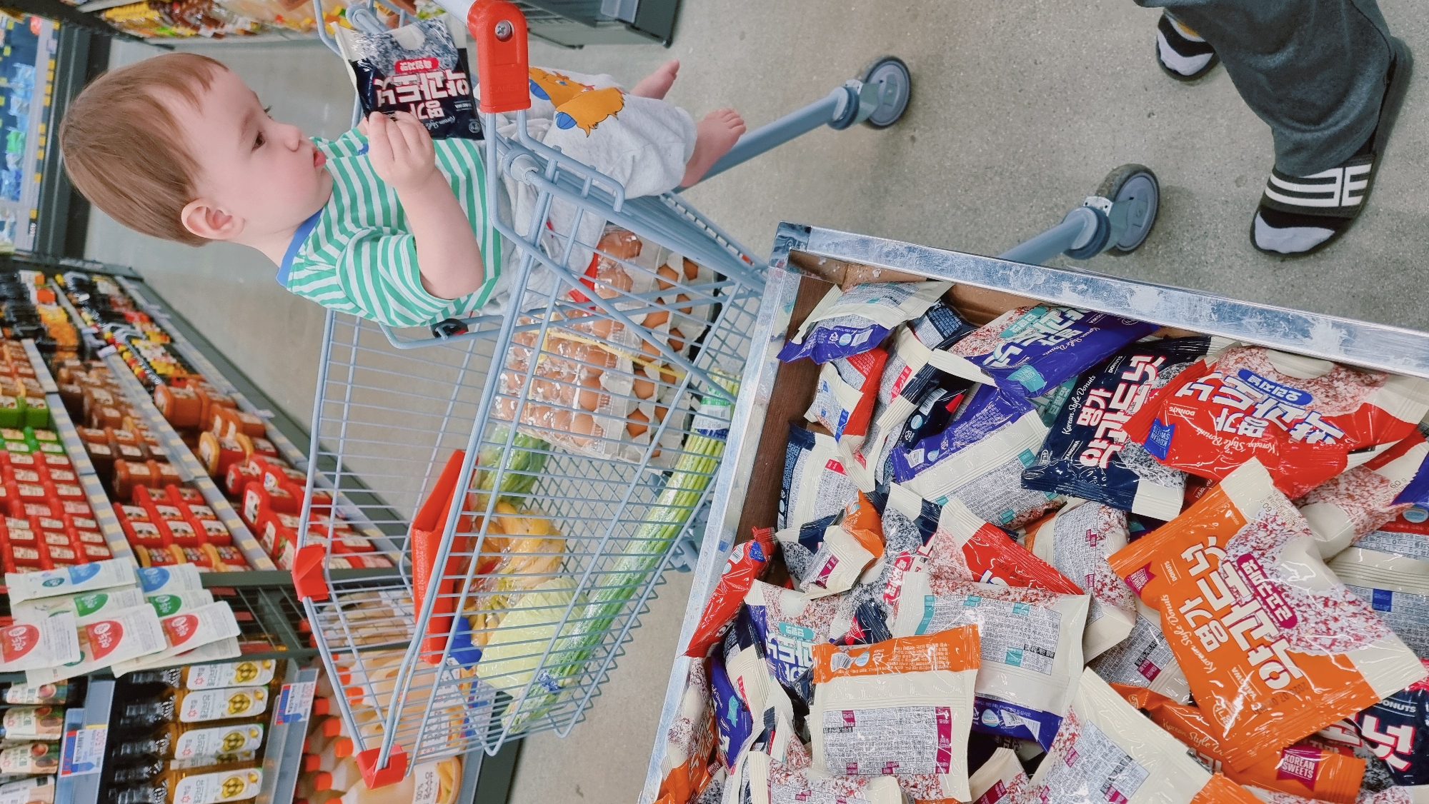 The image depicts the interior of a store in Dunnae-myeon, South Korea. A baby with light brown hair, wearing a green and white striped shirt and white patterned pants, sits in the upper section of a shopping cart. The child holds a black and white package. The cart also contains a carton of eggs, green vegetables, and yellow fruit. Adjacent to the cart is a display of packaged Korean donuts in various flavors, including orange, red, and blue packaging with Korean text. The donuts are labeled as "Korean Style Donuts." Shelves with assorted food products are visible in the background. A person wearing grey pants and black and white slippers stands near the cart. The floor is grey and the lighting is bright.