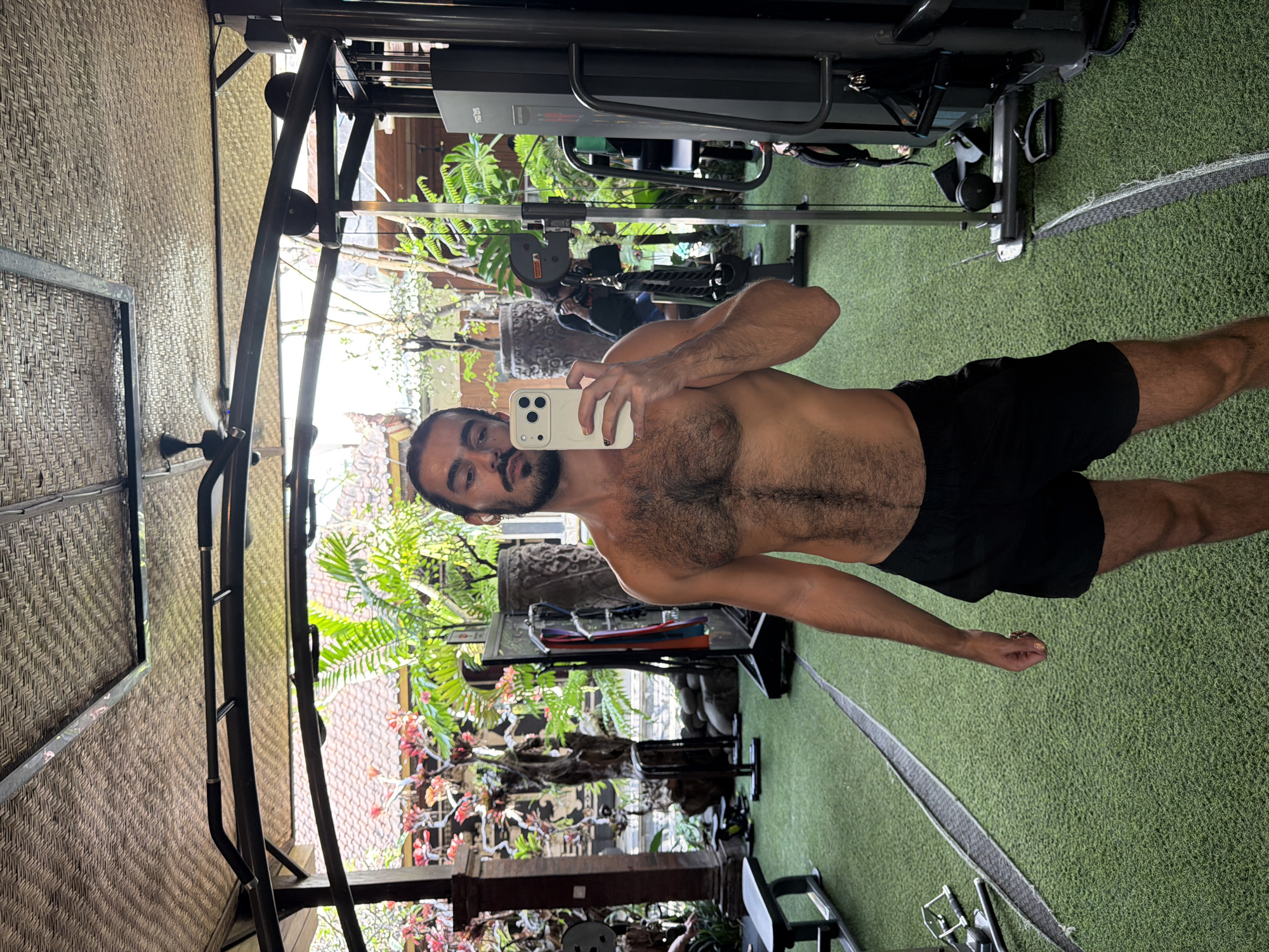 A man with dark hair and a beard takes a selfie in a gym in North Kuta, Indonesia. He is shirtless and wearing black shorts. The gym has artificial turf flooring and is open to the outdoors, with lush greenery visible in the background. There is gym equipment, including weight machines and benches, as well as a ceiling fan. Another person can be seen in the reflection of a mirror in the background.