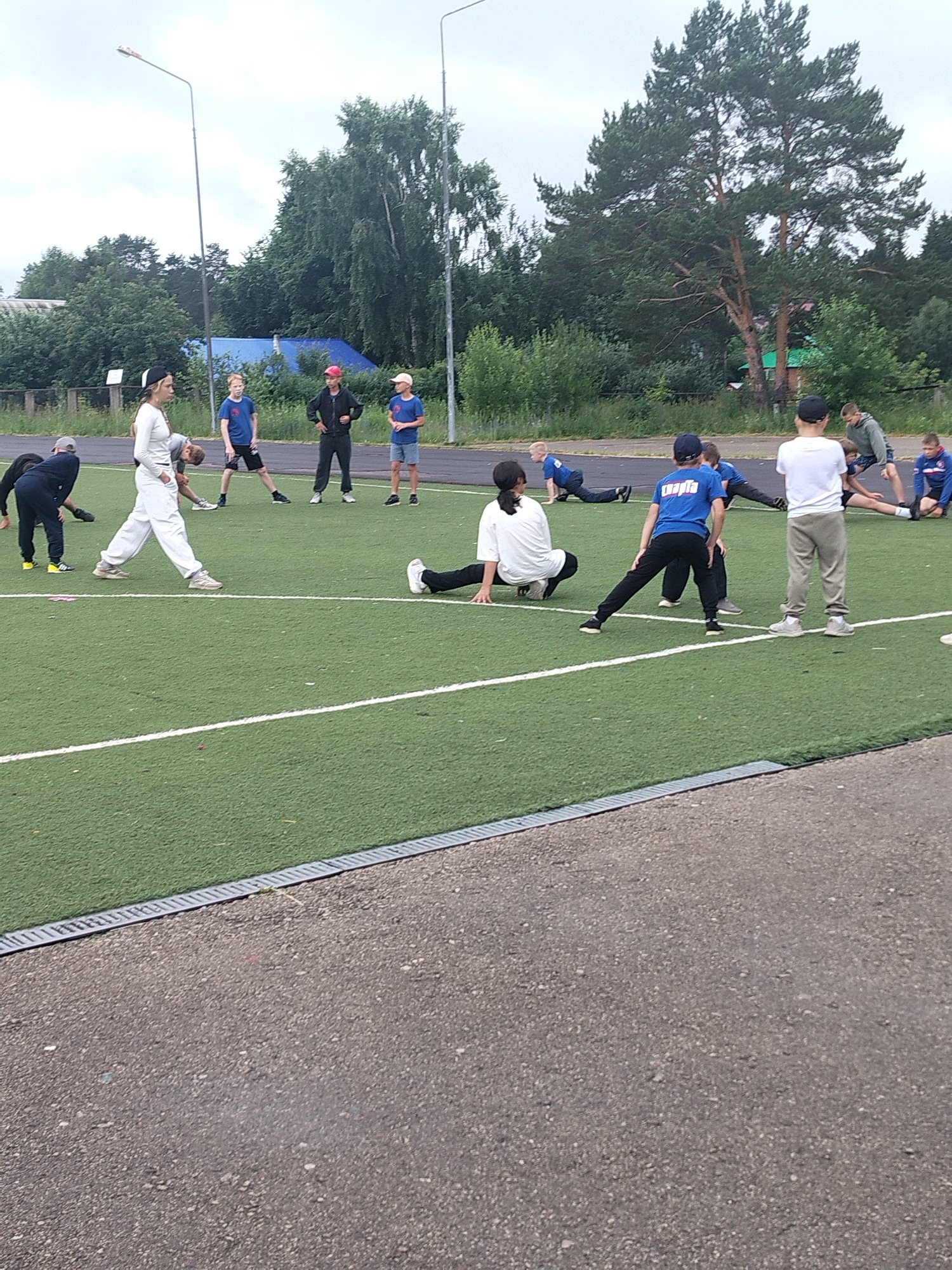 The image depicts an outdoor group training session on a green turf field, likely a sports ground. Several individuals, appearing to be young people, are engaged in stretching or warm-up exercises.
In the foreground, a person wearing white pants and a white top is stretching. To their right, another person in white is also stretching. Further into the field, a group of individuals in blue shirts and black pants are performing similar exercises. One individual in a blue shirt has the word "CAPTAIN" printed on the back. Several other individuals in various athletic wear are scattered across the field, some in motion and others in static stretches. The field is marked with white lines.
The background shows a line of trees and a cloudy sky. In the far distance, a blue structure and what appears to be a street lamp are visible. The ground bordering the field is dark gray asphalt. The overall environment suggests a public sports facility.
The presence of a "CAPTAIN" shirt and the organized activity implies a sports team or a structured training event. The setting is a moderately green, open area typical of an athletic field. The clothing of the participants suggests a casual, active setting.
Without further context or specific landmarks within the image, pinpointing the exact location in Chaikovsky, Russia is not possible. However, the scene is consistent with a typical sports ground found in many urban or suburban areas.