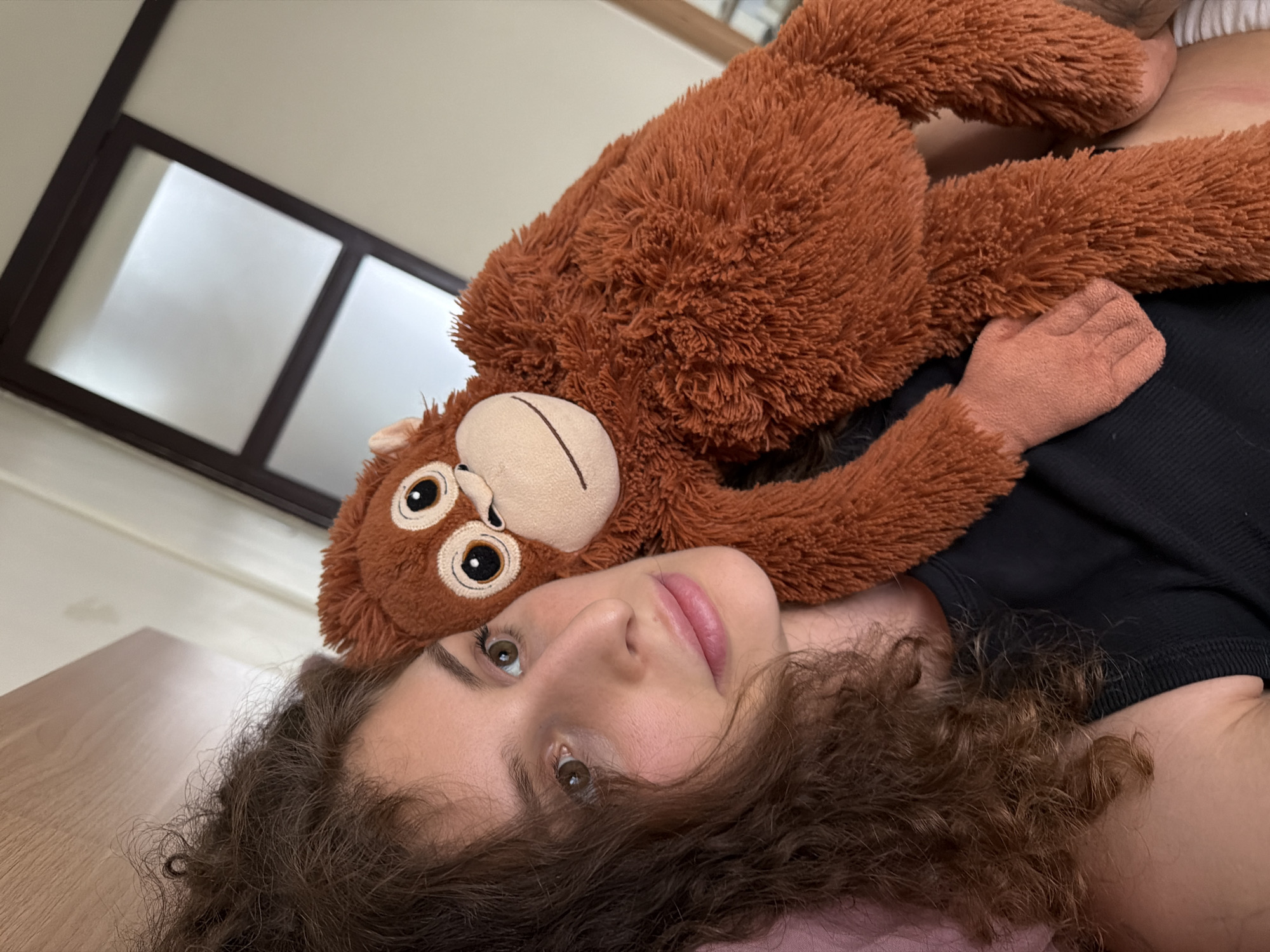 A young woman with curly brown hair is lying down, holding a stuffed orangutan. She is wearing a black top and her face is partially obscured by her hair. The orangutan is brown and has large, round eyes. They are in a room with a wooden headboard behind them and a window with a dark frame in the background. The room appears to be a bedroom. The scene is captured from a close-up, high-angle perspective.