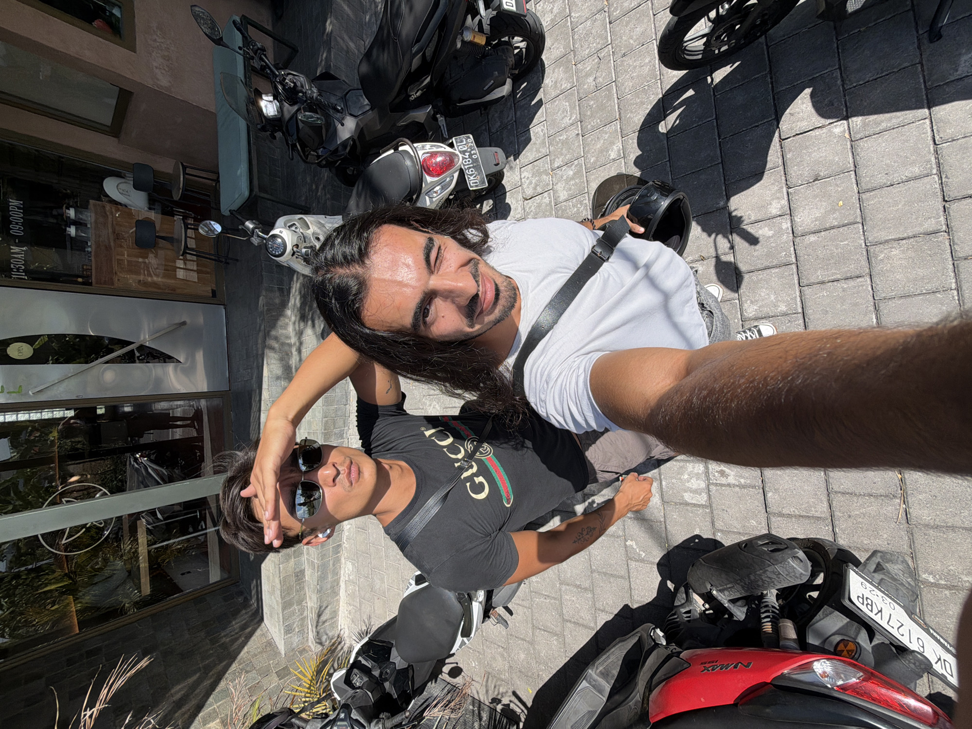 The image, oriented in a selfie-style overhead shot, shows two men standing among parked motorbikes on a brick-paved surface. The taller man, with long dark hair, wears a white shirt with a black strap across his chest. He is positioned on the right side of the frame, smiling at the camera, holding a black helmet in his left hand.
The second man, slightly shorter, stands to the left of the frame. He is wearing sunglasses and a black Gucci t-shirt. His right hand touches the top of his head.
Several motorbikes are parked around the men, positioned at the top and bottom edges of the frame. At the top, one motorbike has a license plate reading "DK6184.0L". At the bottom, another motorbike has a license plate reading "DK 6127 KBP".
In the background, a shop window with a sign indicating "OPEN 11:30AM - 09:00PM" is visible, suggesting the location is North Kuta, Indonesia.
