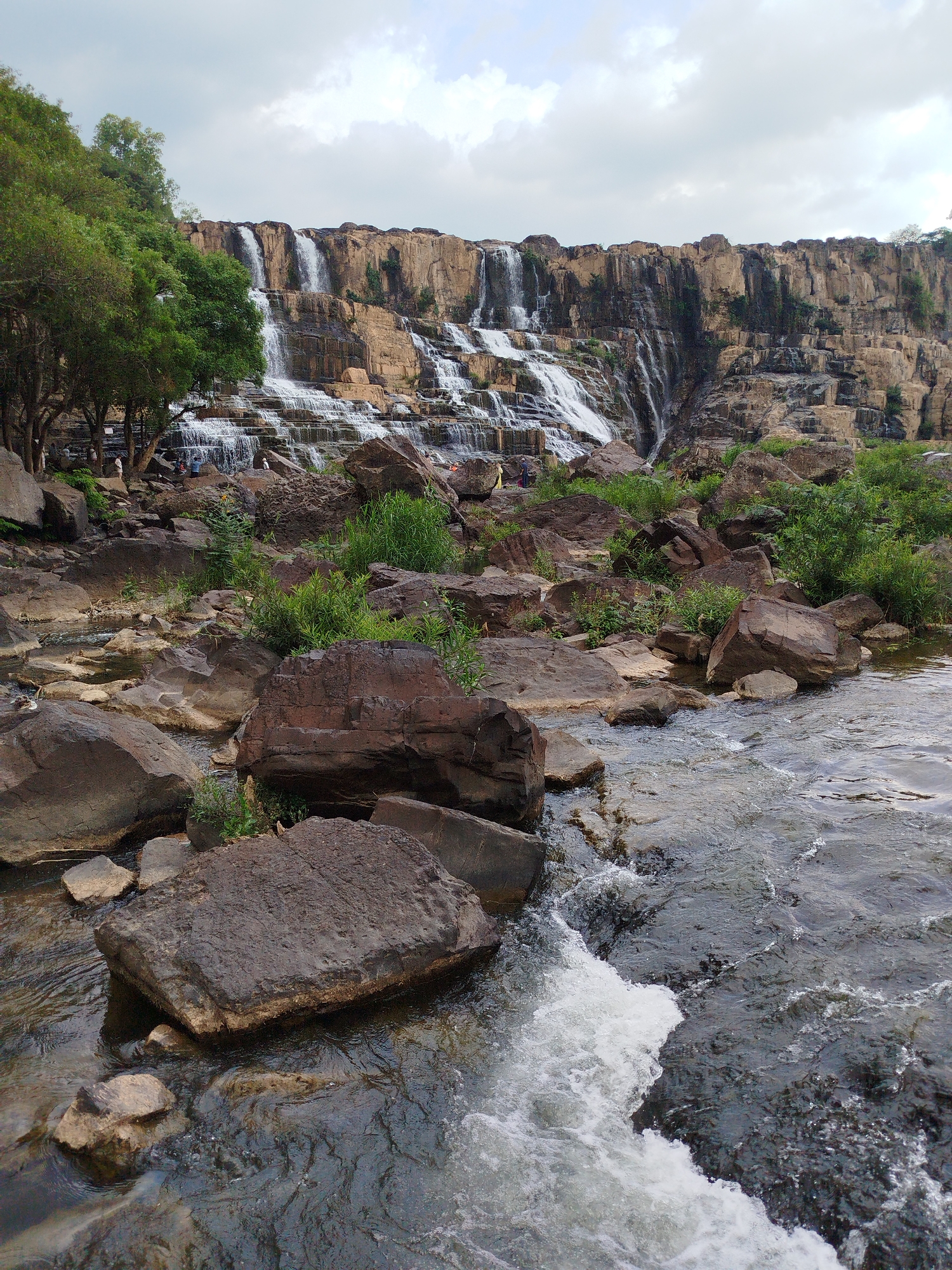 The image shows a waterfall landscape in Tân Phú, Vietnam, under a cloudy sky. The falls cascade down a multi-tiered rock face. The foreground features a stream with water flowing around large, dark-colored rocks. Green vegetation grows among the rocks. In the midground, trees are visible. People appear near the base of the waterfall. The scene conveys a natural environment with a combination of rock, water, and greenery.