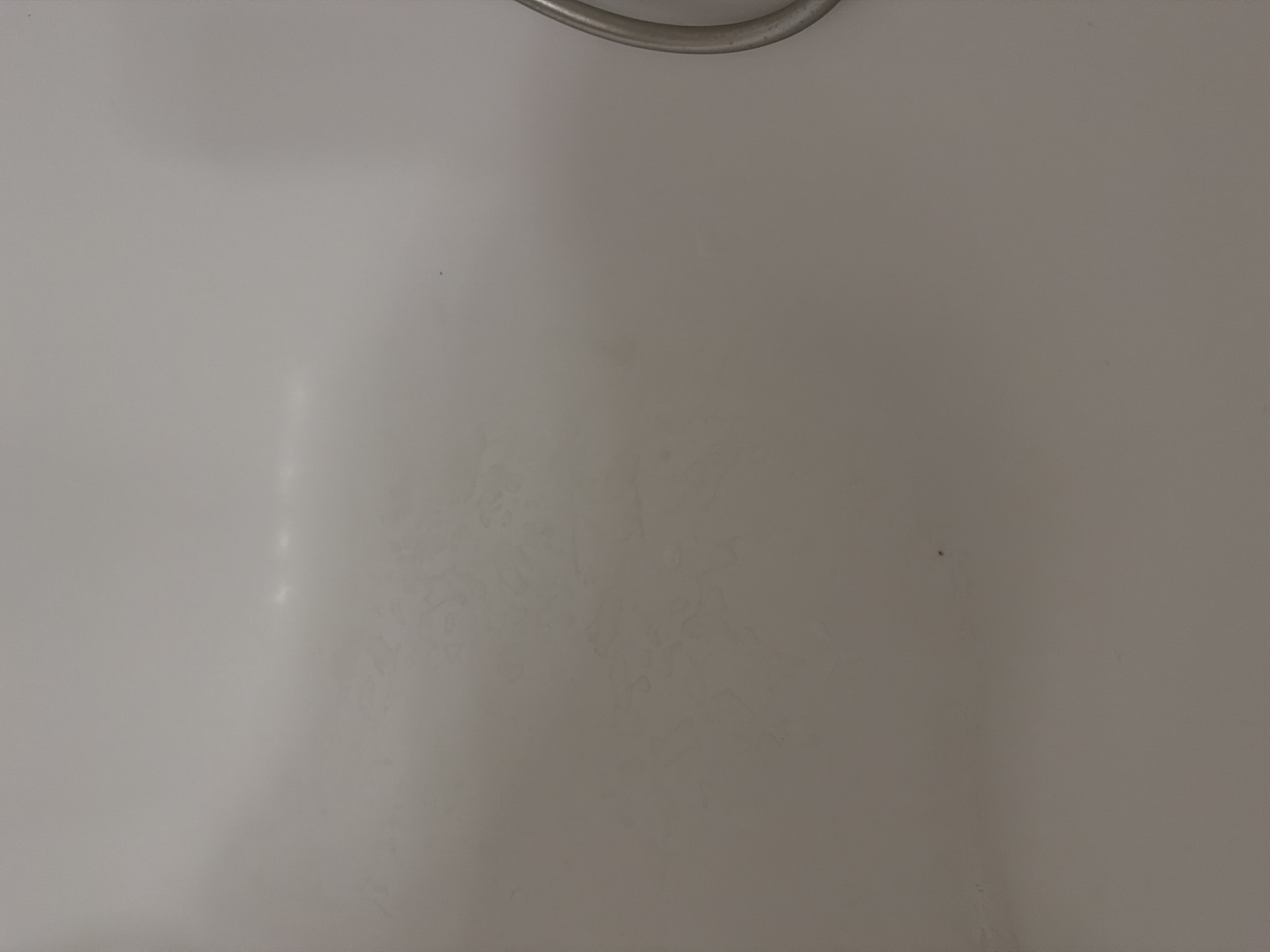 The image is a close-up, overhead shot of a dull, light-colored surface, possibly a wall or a countertop. The lighting is soft, with a faint horizontal reflection visible near the top center of the frame. There are subtle, irregular patterns and smudges on the surface, indicating it may not be perfectly clean. In the far right of the image, a portion of a silver, circular object is visible, possibly a handle or a ring. The image is devoid of any identifiable landmarks or features that would allow for its specific location within Khmelnytskyi, Ukraine to be determined.