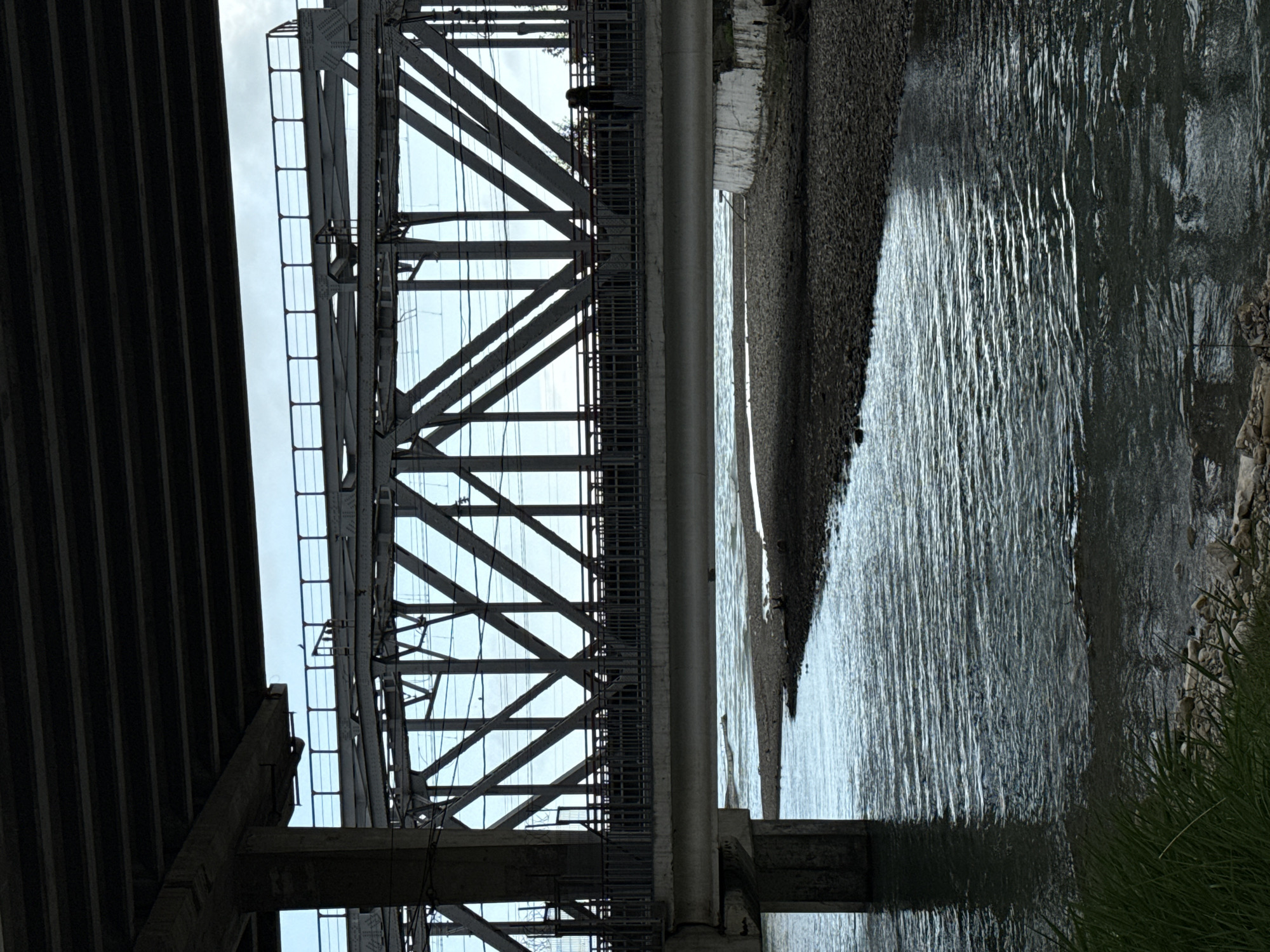 A low-angle, medium shot photograph depicts a structural bridge, possibly a train or vehicle bridge, crossing over a body of water. The bridge's support structure is made of gray metal girders arranged in a diagonal, crisscross pattern. The bridge is seen from underneath, looking upwards towards the sky, which is visible through the steel framework. A lower deck of the bridge is visible on the left side of the frame.
On the right, the body of water reflects the light and bridge structure. A narrow, sandy or pebbled shore borders the water, and vegetation is seen growing on the banks. The lighting is overcast, casting a soft, diffused light on the scene. The location appears to be somewhere in the city of Unknown, Unknown.