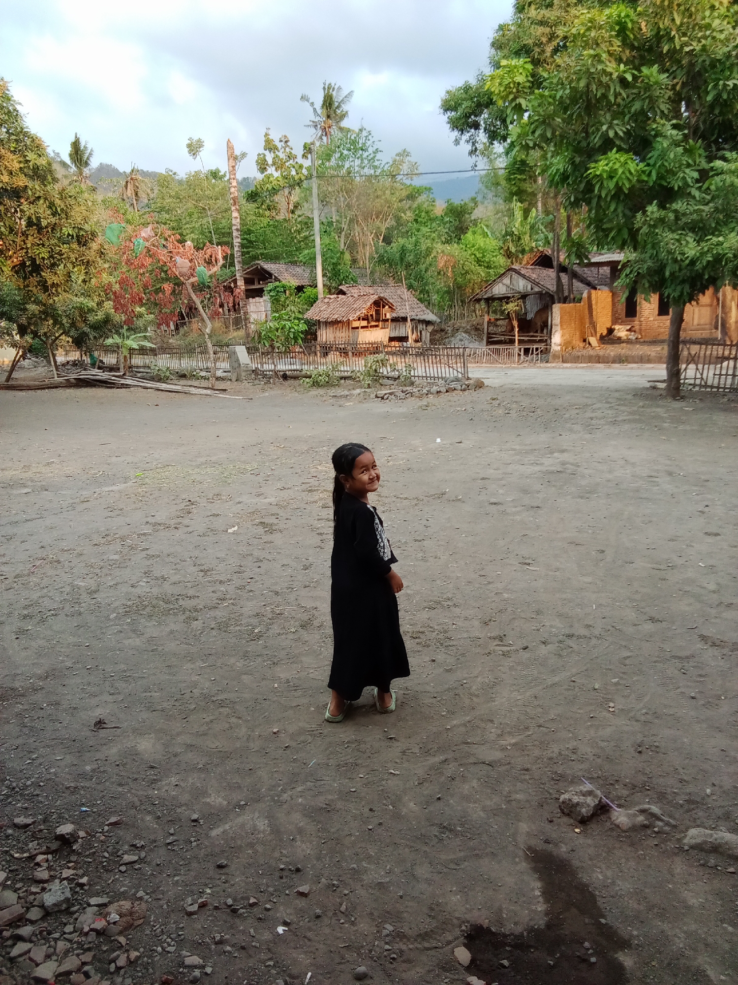 In a rural village setting in Selowogo, Indonesia, a young girl dressed in a black long-sleeved dress and light-colored shoes stands facing to her right, looking back towards the camera with a smile. She has dark hair styled in a braid. The ground is a dusty, uneven dirt surface with scattered rocks and debris. In the background, traditional Indonesian village structures with thatched roofs are visible, surrounded by lush green trees and vegetation. The sky is overcast, suggesting it may be early morning or late afternoon. The scene depicts a slice of everyday life in a rural Indonesian community.
