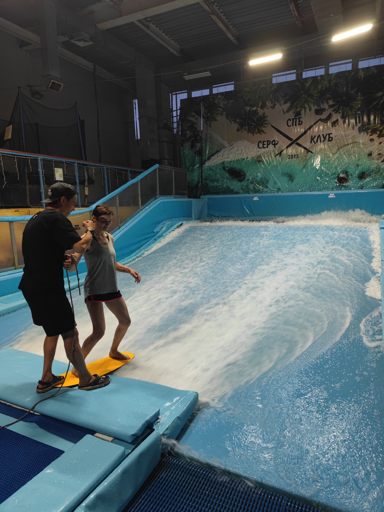 This media file captures an indoor surfing activity at a surf club in Saint Petersburg, Russia. The scene features two people: an instructor, a male wearing a black t-shirt and shorts, and a participant, a female wearing a grey tank top and dark shorts with a colorful trim. The instructor is guiding the participant who is standing on a yellow board, preparing to surf on a simulated wave.
The setting is an indoor surf pool with bright blue padding around the edges and a dynamic water surface creating a wave. The background shows a large, colorful mural depicting a tropical beach scene with palm trees, sand, and water. The mural also features text that reads "СПБ СЕРФ КЛУБ 2015" along with two crossed surfboards. The lighting suggests an indoor environment, possibly during the day or evening, with artificial lights visible on the ceiling. The overall mood appears to be active and focused, with the participant concentrating on the surfing lesson. The presence of safety equipment like the blue padding and the instructor's guidance indicates a controlled and instructional environment. The water is visibly churning and creating a wave, highlighting the core activity of the surf club.