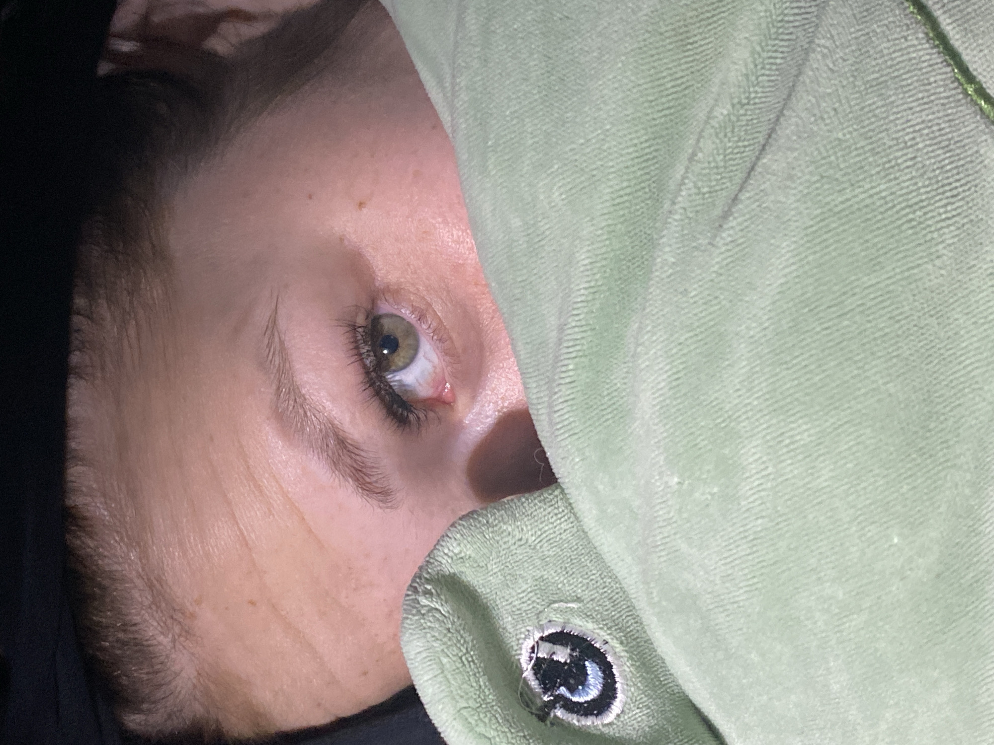 The image shows a person partially obscured by a light green fabric, likely a blanket or hoodie. Only one eye is fully visible; it's hazel in color. Part of the face and hairline are also visible. The fabric has a logo or patch visible in the bottom center. The environment is dark. The location in the city of Chaikovsky, Russia, is unknown.