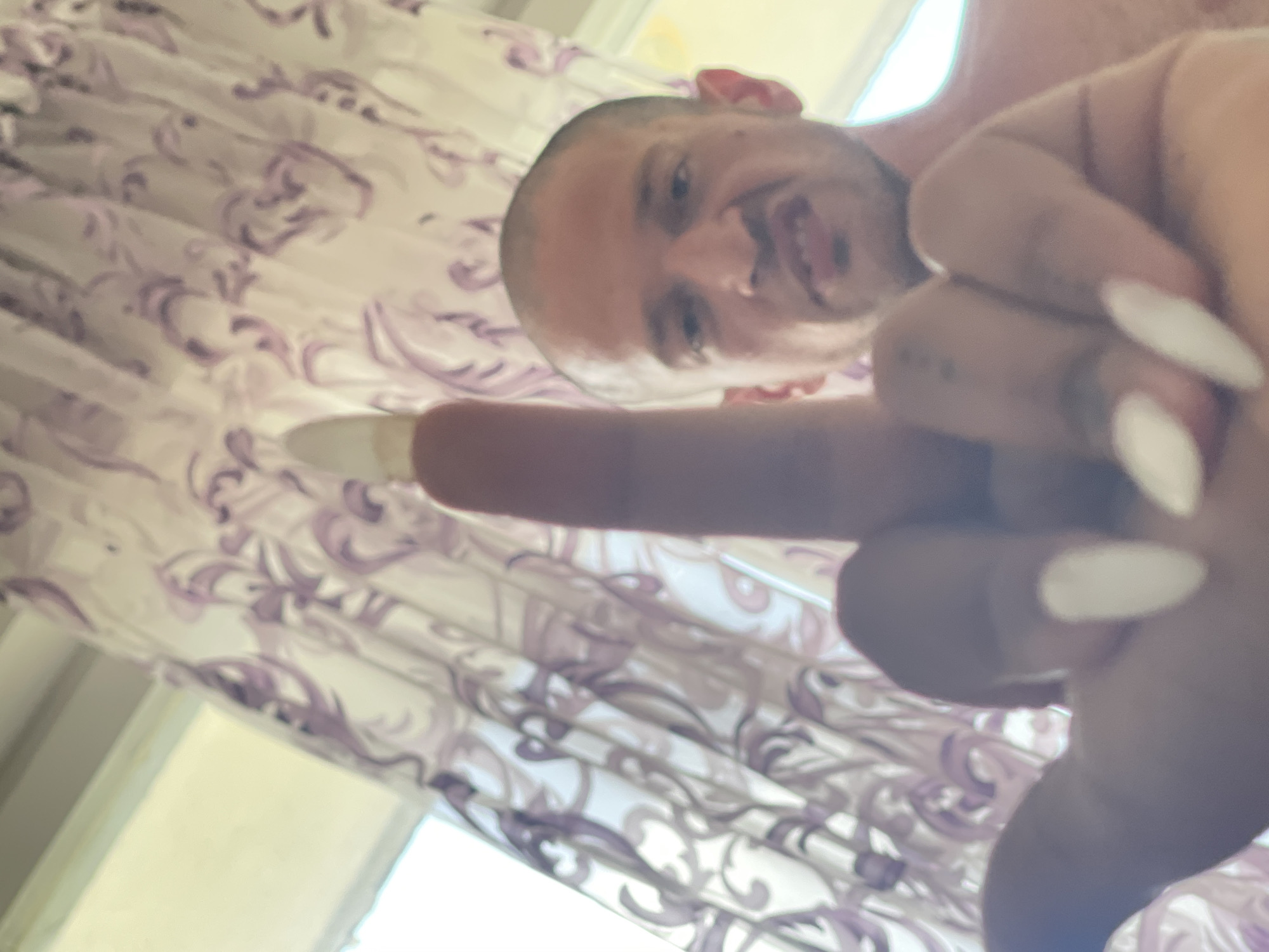 A close-up, low-angle shot shows a person's hand with long, white, pointed fingernails extended towards the camera, with the middle finger raised in a vulgar gesture. Behind the hand, a man with a shaved head and a scowling expression is visible, looking directly at the camera. The background consists of light-colored curtains with a purple floral pattern. The image appears to have been taken indoors, possibly in a residential setting. There is no information available regarding the location within Varna, Bulgaria.