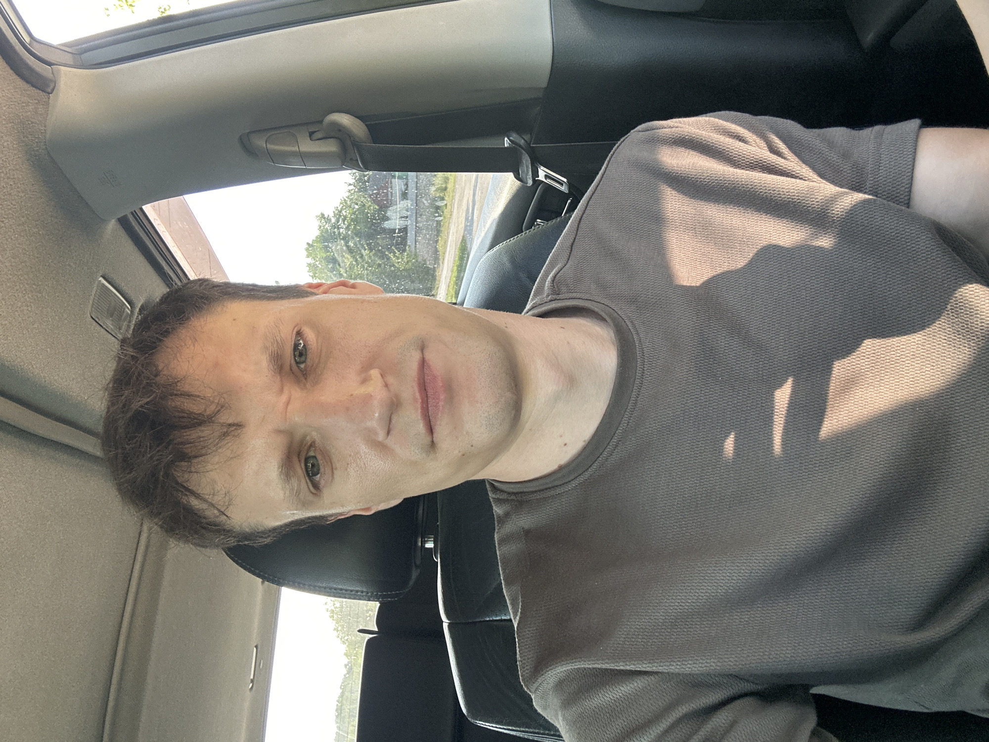 A close-up, waist-up selfie of a fair-skinned male with short brown hair is captured inside a car. He is looking directly at the camera with a neutral expression. The man is wearing a brown t-shirt with a textured waffle knit pattern. Sunlight casts shadows across his shirt and face, indicating it is daytime. The interior of the car is visible, including black leather seats, the ceiling lining, and a grey pillar with a seatbelt buckle. The background outside the car window shows greenery and a road, suggesting a rural or suburban setting, possibly on the outskirts of Makiv, Ukraine.
